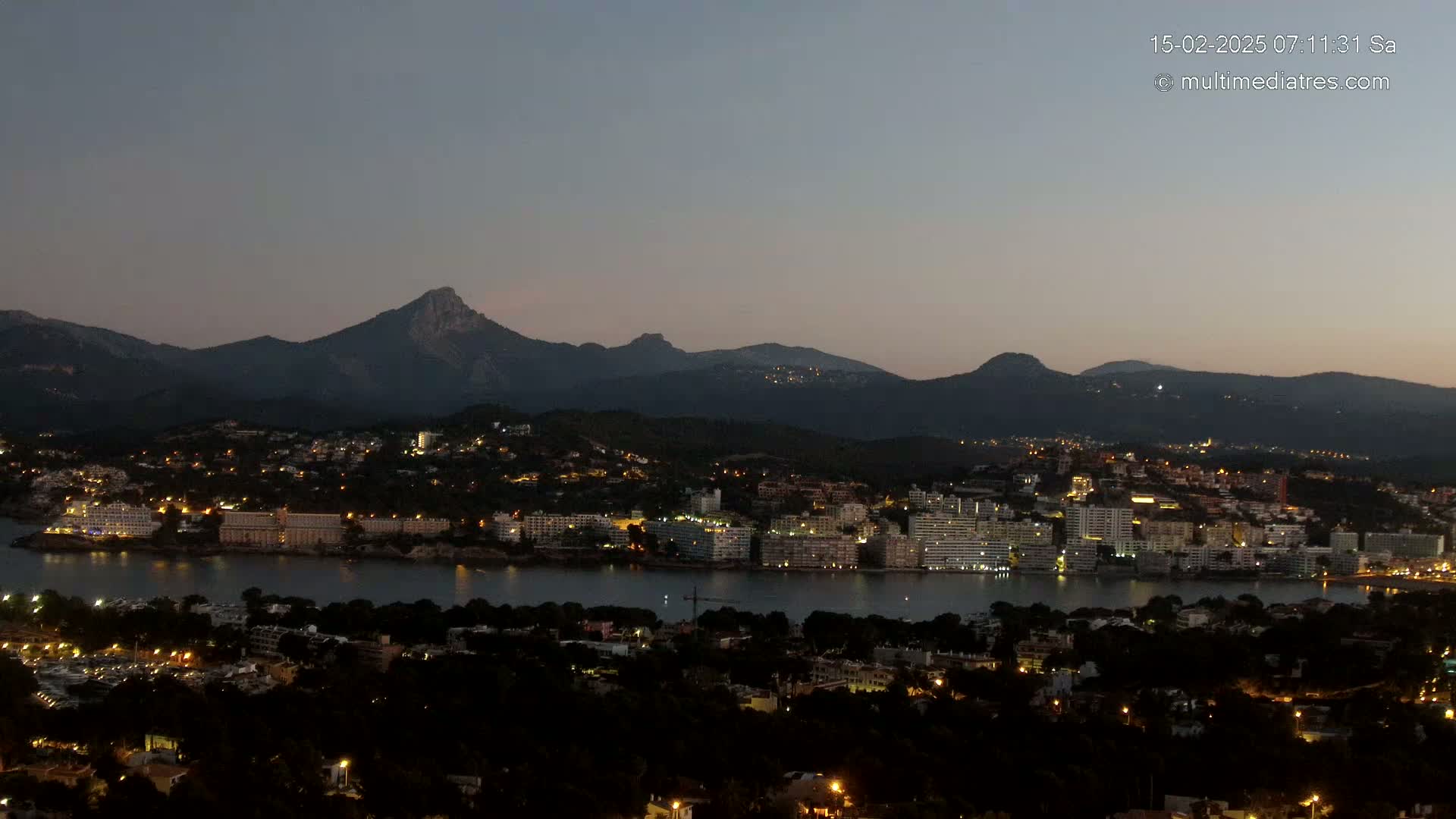 A coastal town at night, illuminated by lights, is nestled below a range of dark mountains under a twilight sky.