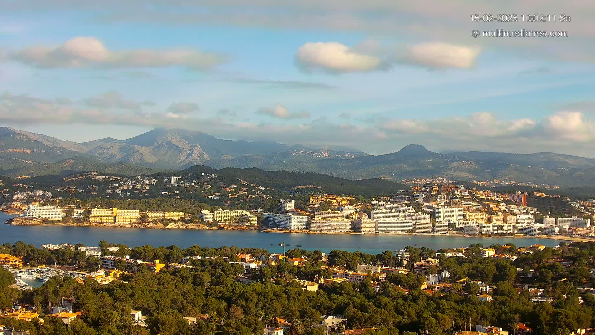 A coastal town nestled beside a calm bay, with a mountainous backdrop under a partly cloudy sky.