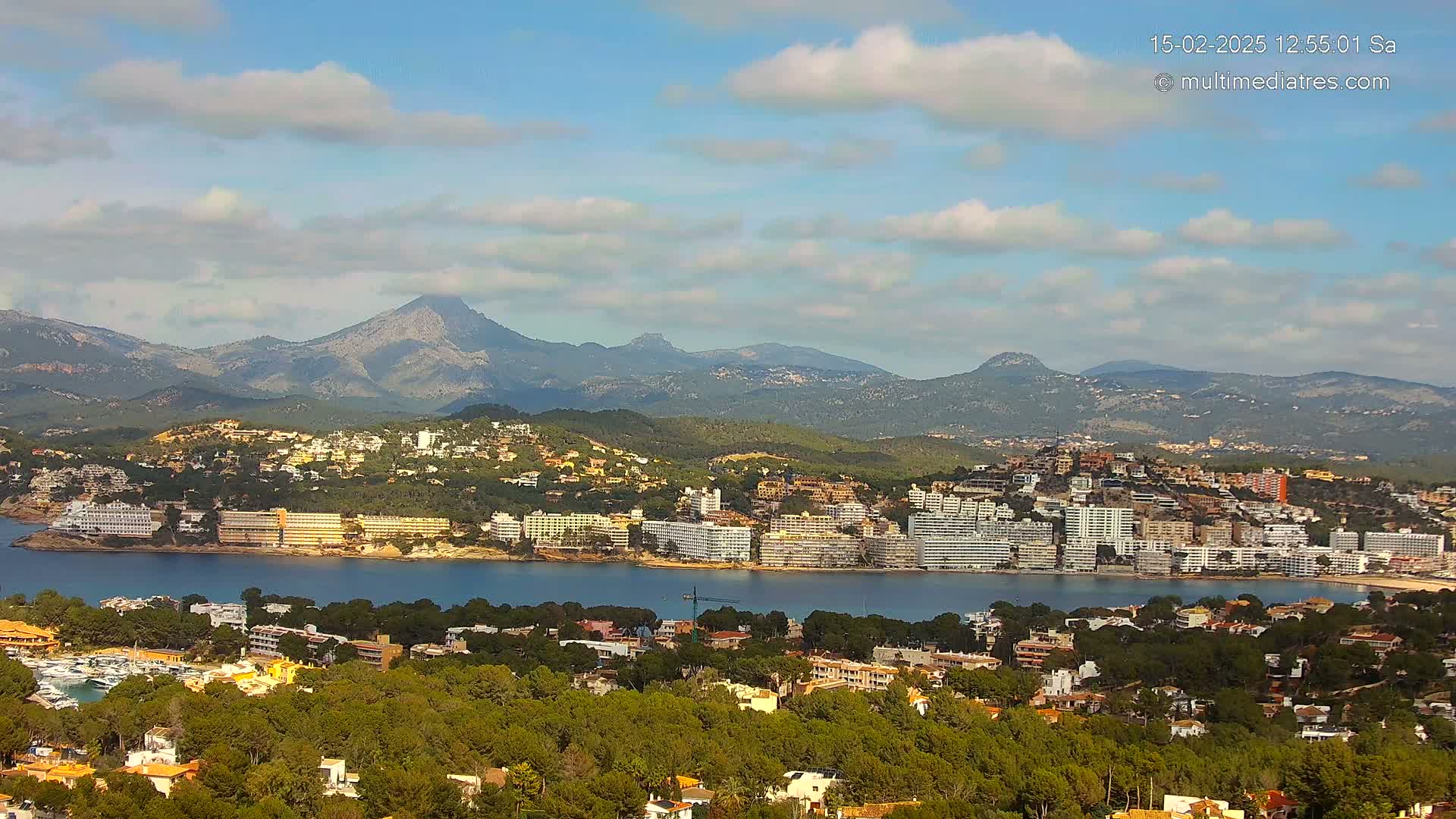 A coastal town nestled amongst green hills and mountains under a partly cloudy sky.
