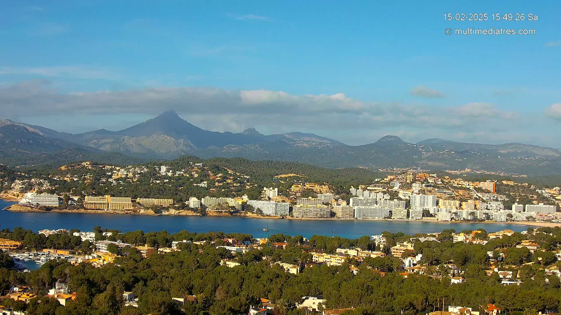 A coastal town with numerous hotels and buildings nestled among green hills, overlooks a calm bay under a mostly clear, sunny sky with some clouds.