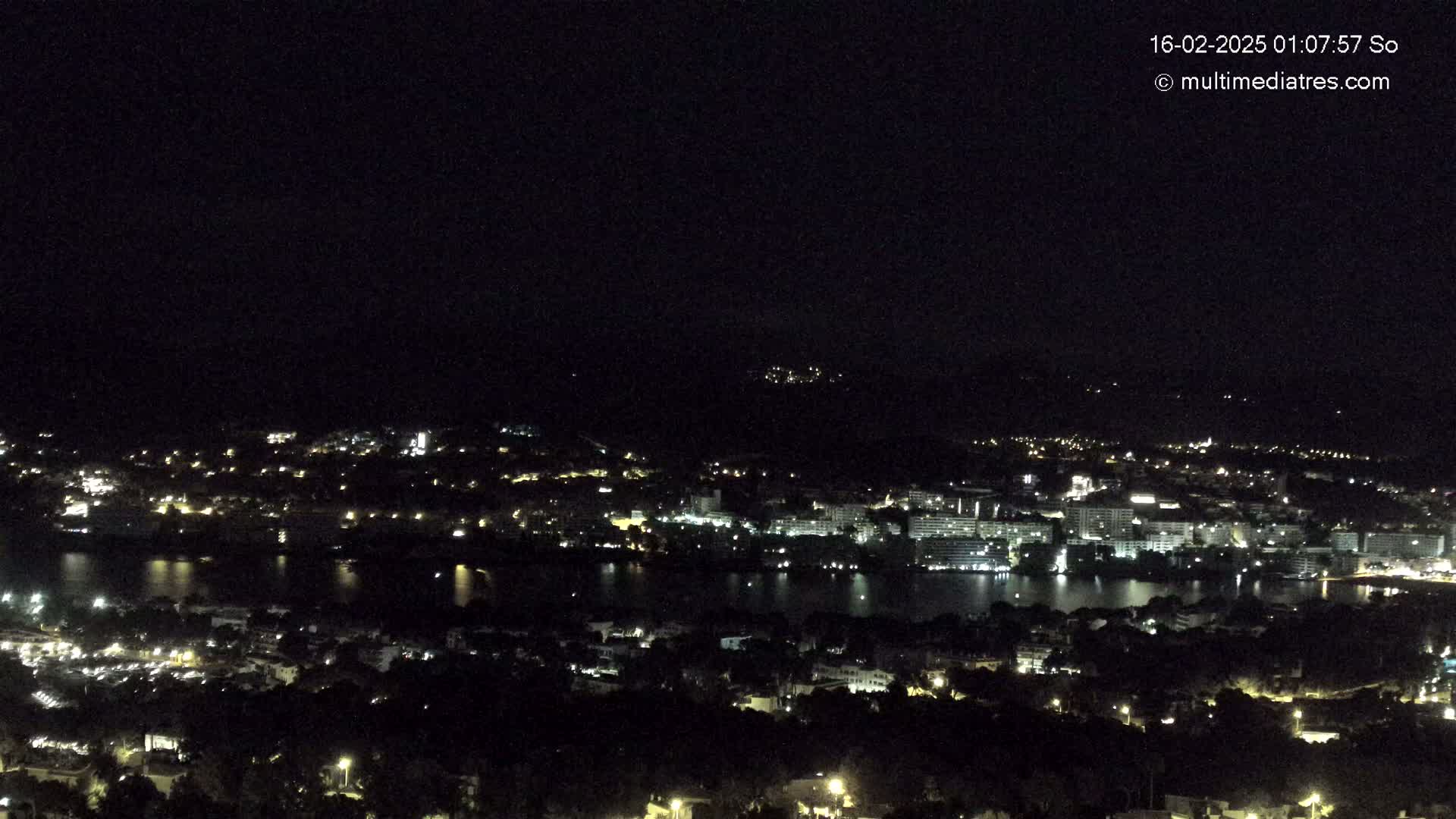 A nighttime, high-angle view shows a coastal city illuminated by scattered lights, with a dark body of water reflecting some of the city lights.