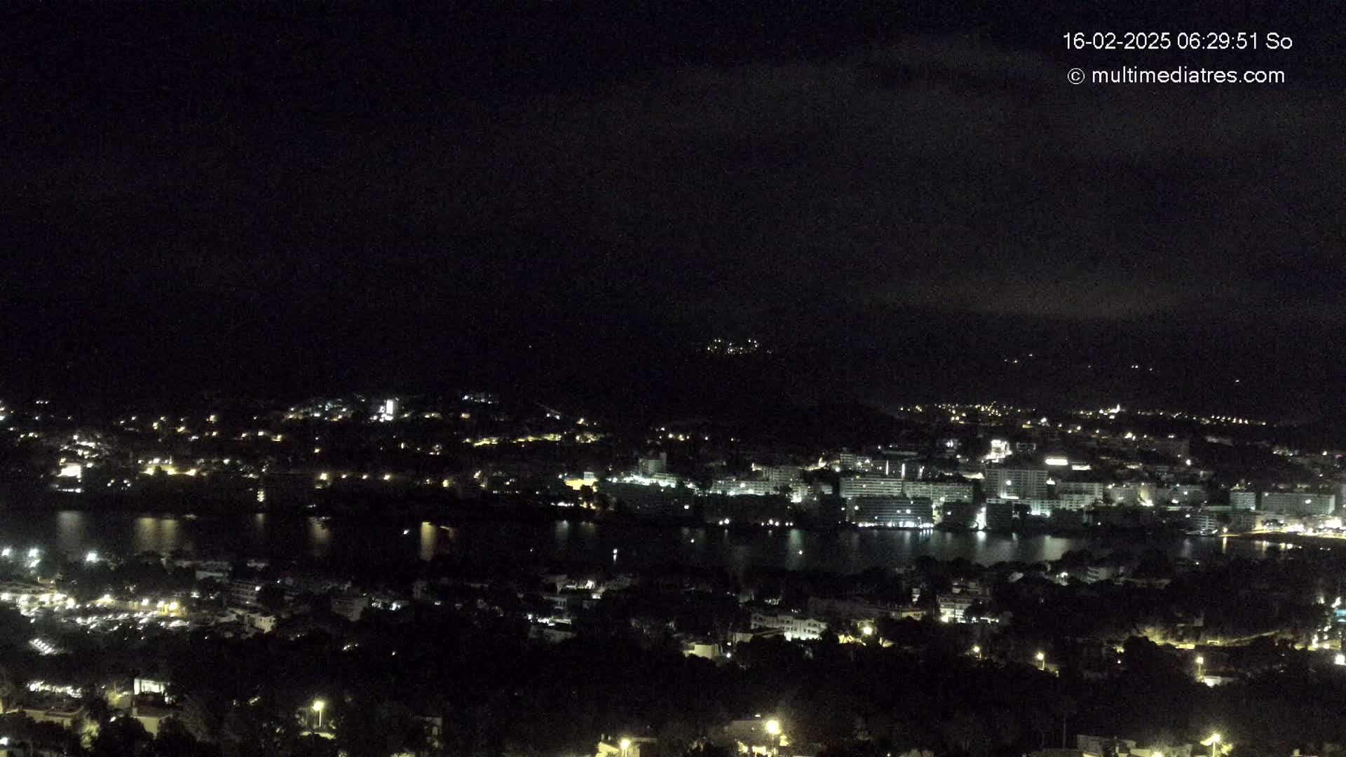 A nighttime view of a city sprawling across a dark body of water, nestled below a hill under a mostly clear sky.