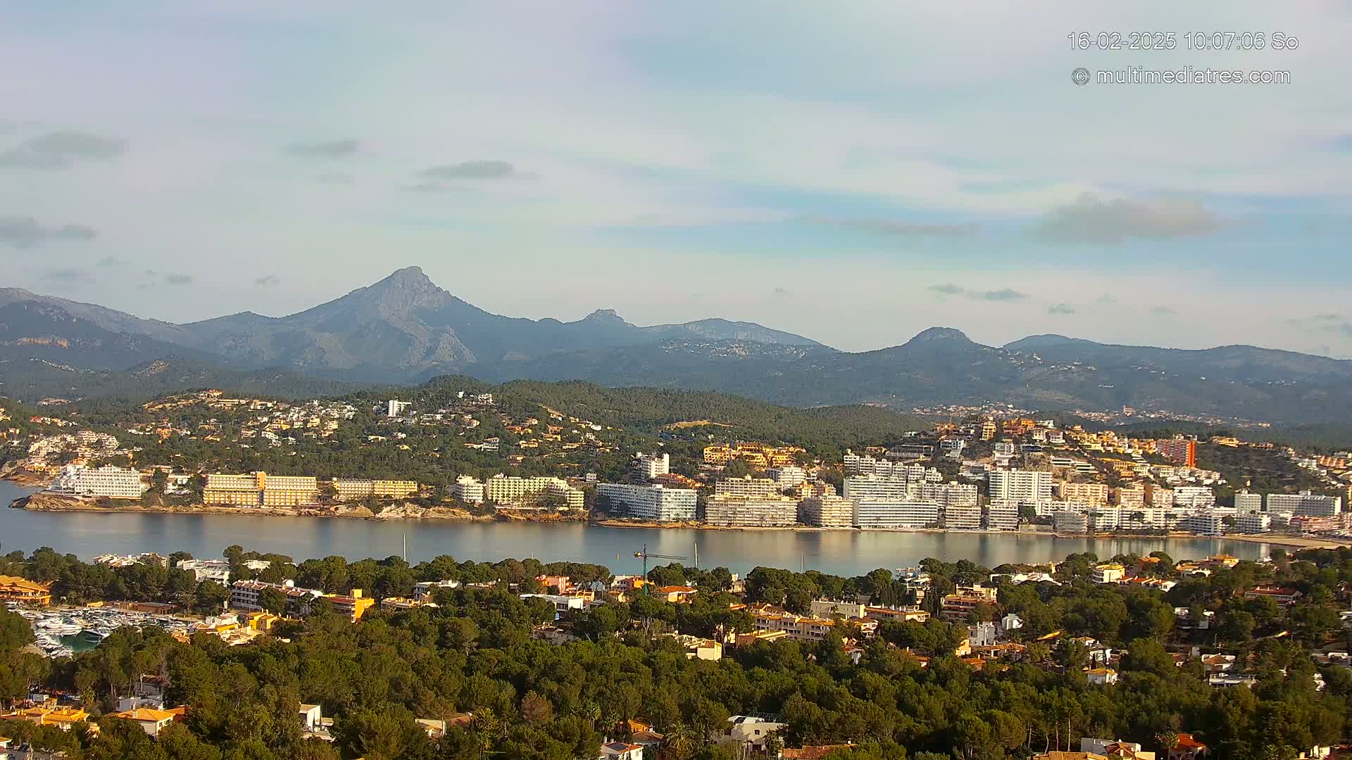 A coastal town nestled between a calm bay and a mountain range under a mostly clear sky.