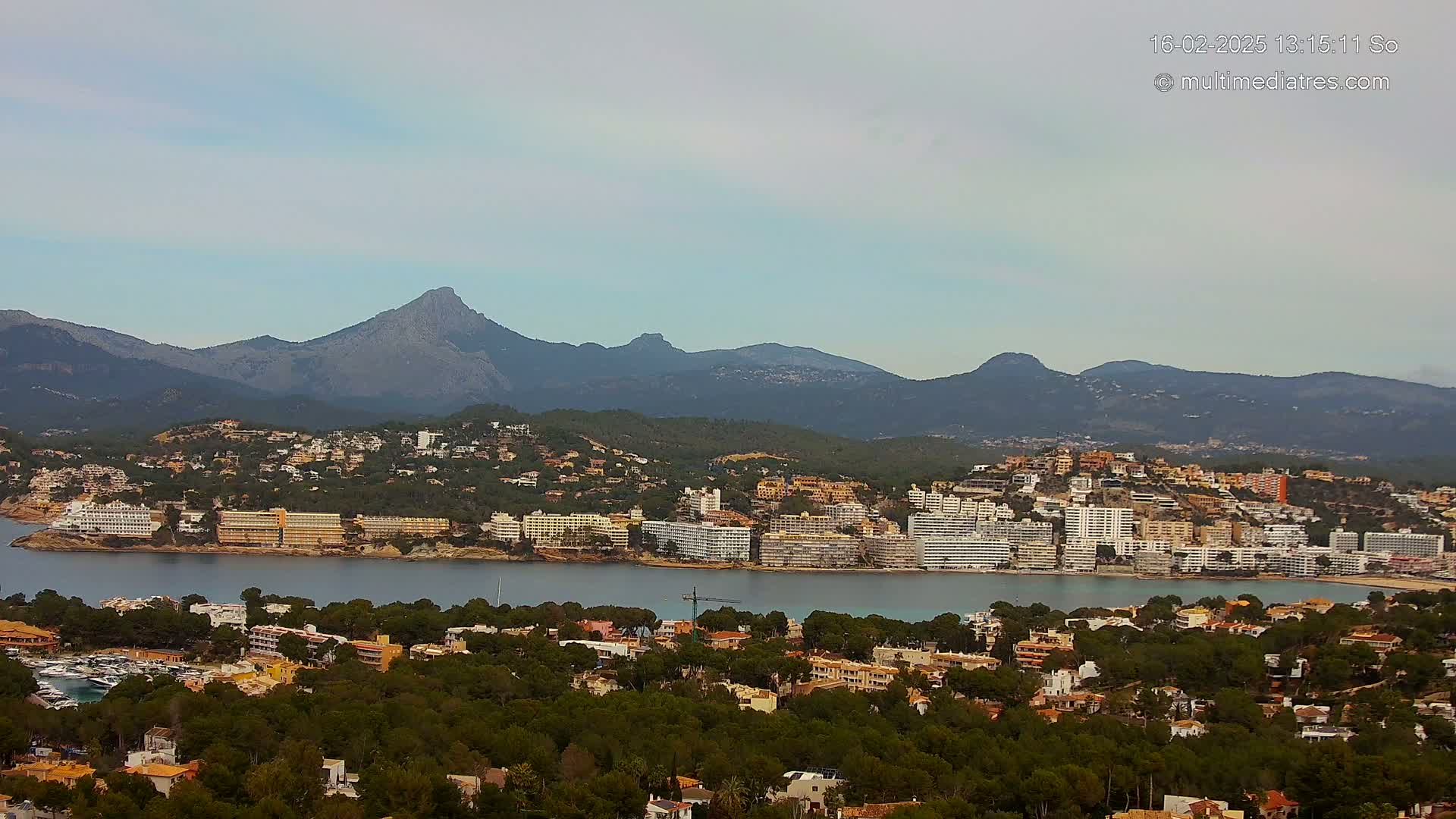A coastal town with numerous buildings sits nestled in a verdant landscape, overlooked by a range of mountains under a mostly clear sky.