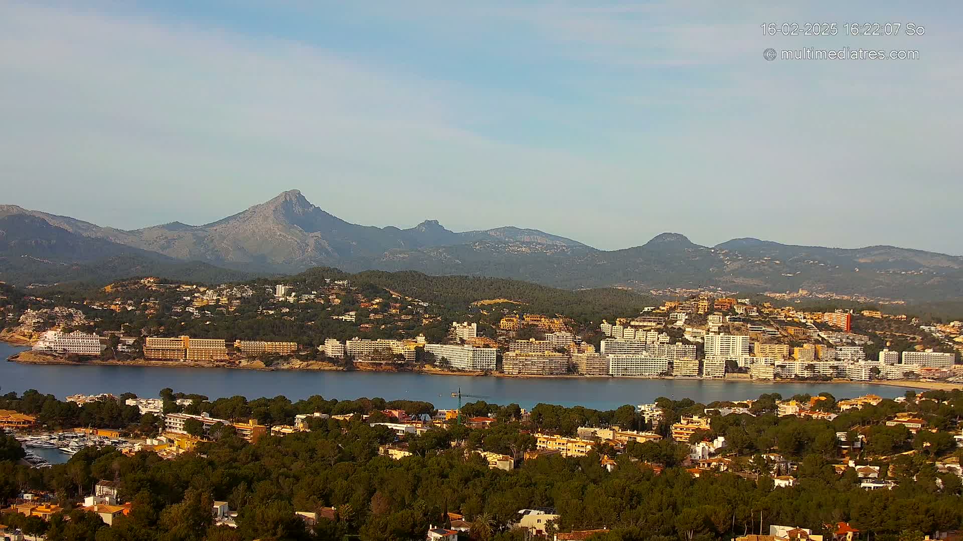 A coastal town nestled among green hills and mountains under a clear sky, with a calm bay separating the town from a line of hotels.