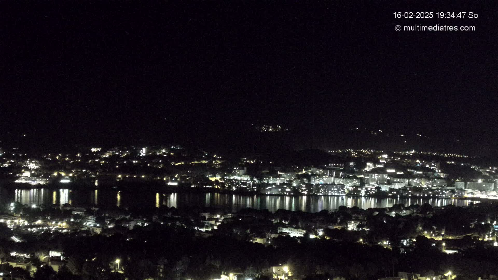 A nighttime view of a city and body of water, illuminated by numerous lights reflecting on the still water under a clear, dark sky.