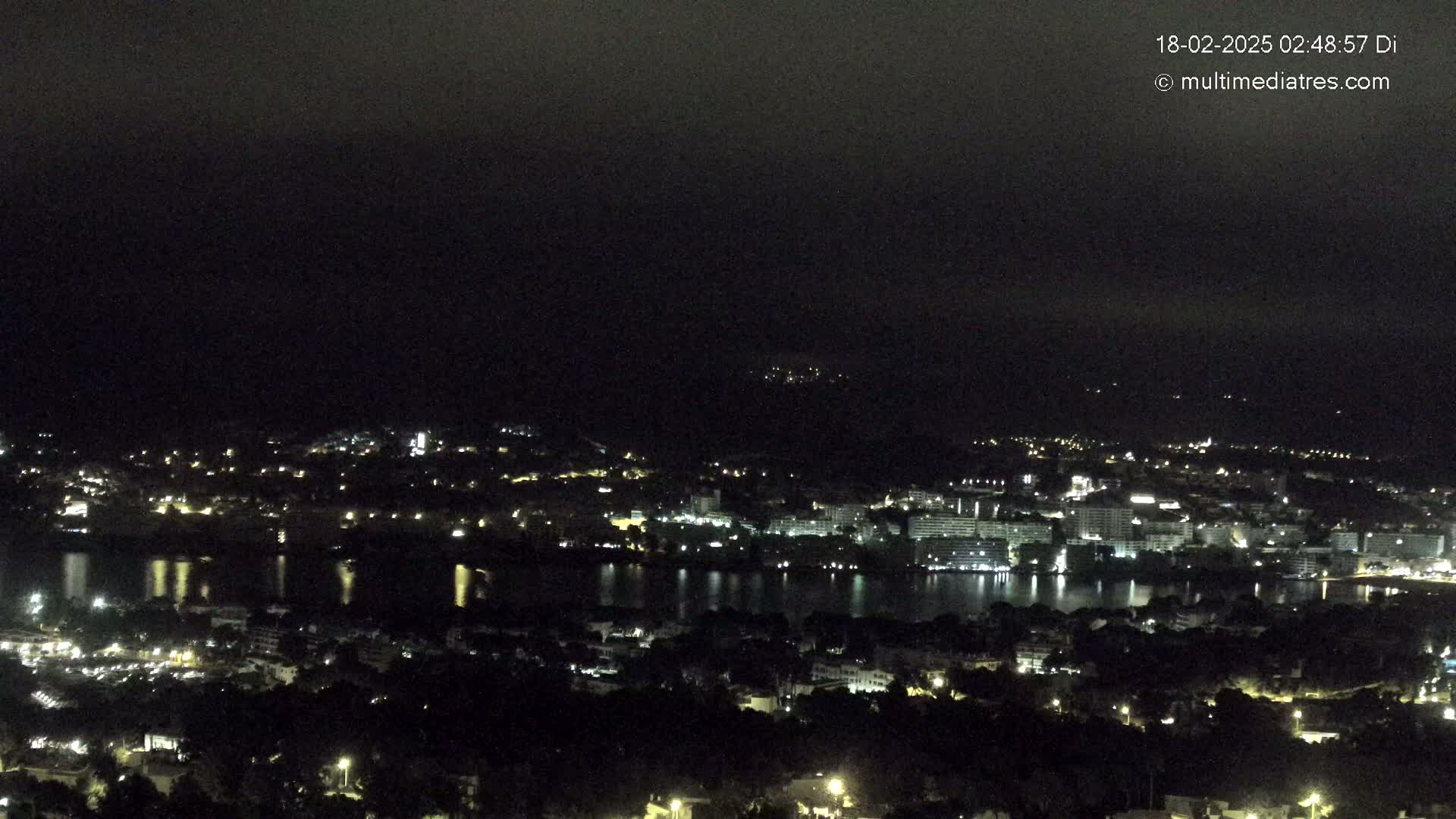 A nighttime, long-range view shows a city with lights illuminating buildings next to a dark body of water, with hills in the background under a dark, cloudy sky.