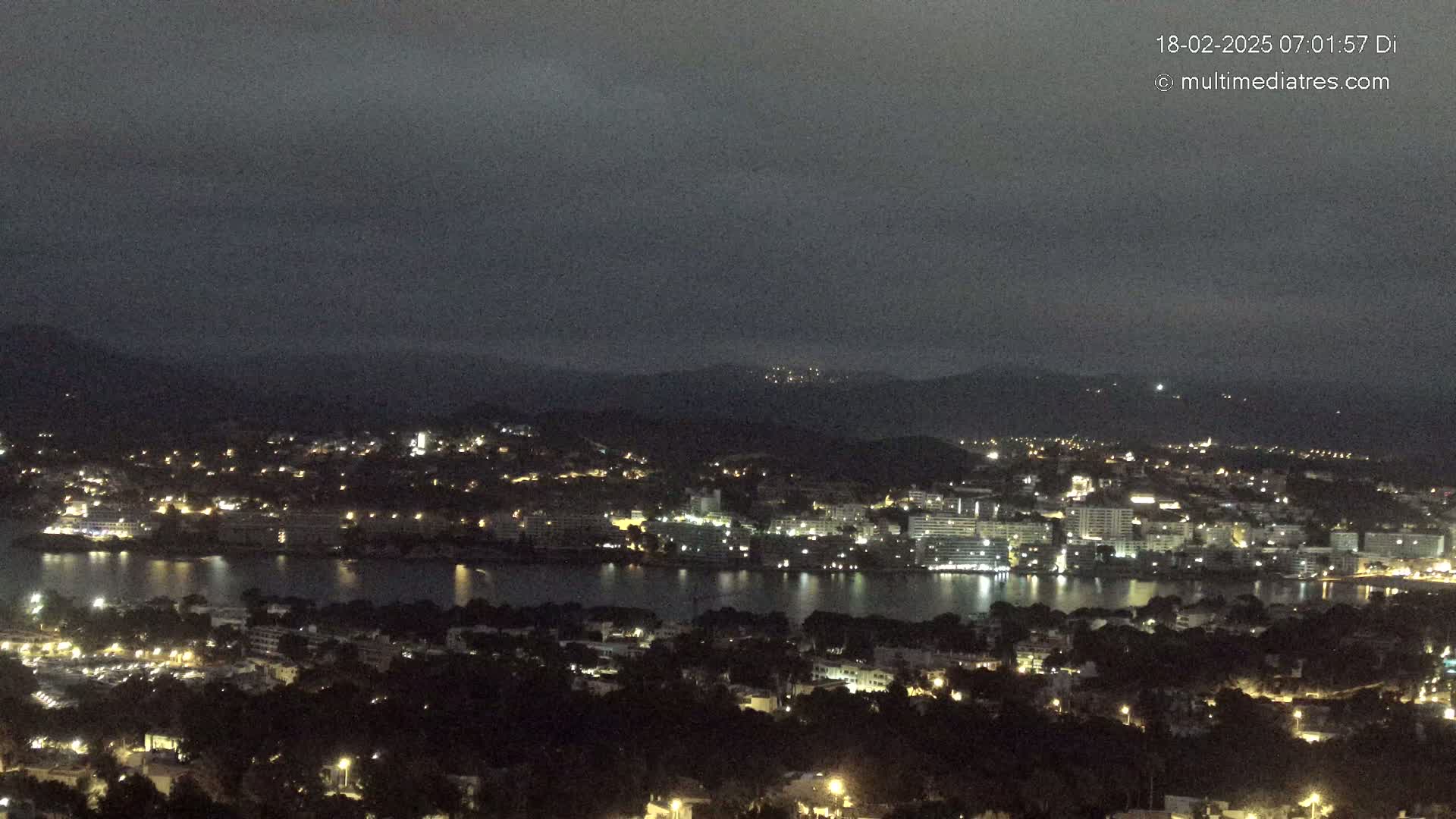 A nighttime view of a coastal town with a body of water, illuminated by scattered lights under a dark, cloudy sky.