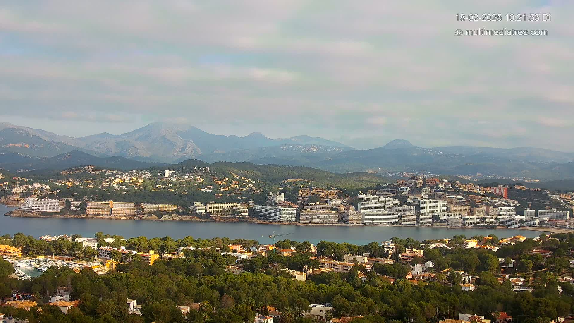 A coastal town nestled beside a calm bay, backed by lush green hills and a hazy mountain range under a mostly cloudy sky.