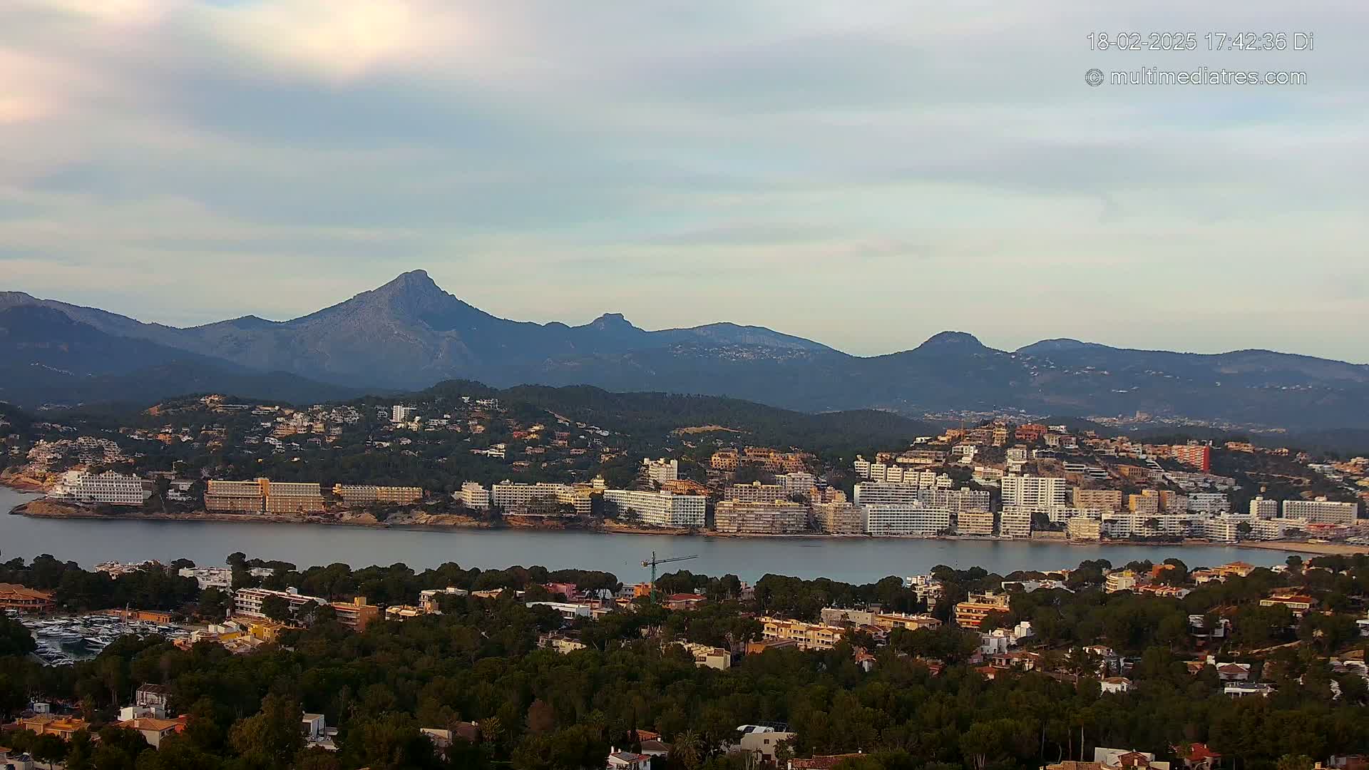 A coastal town nestled between a calm bay and a range of mountains under a partly cloudy sky.