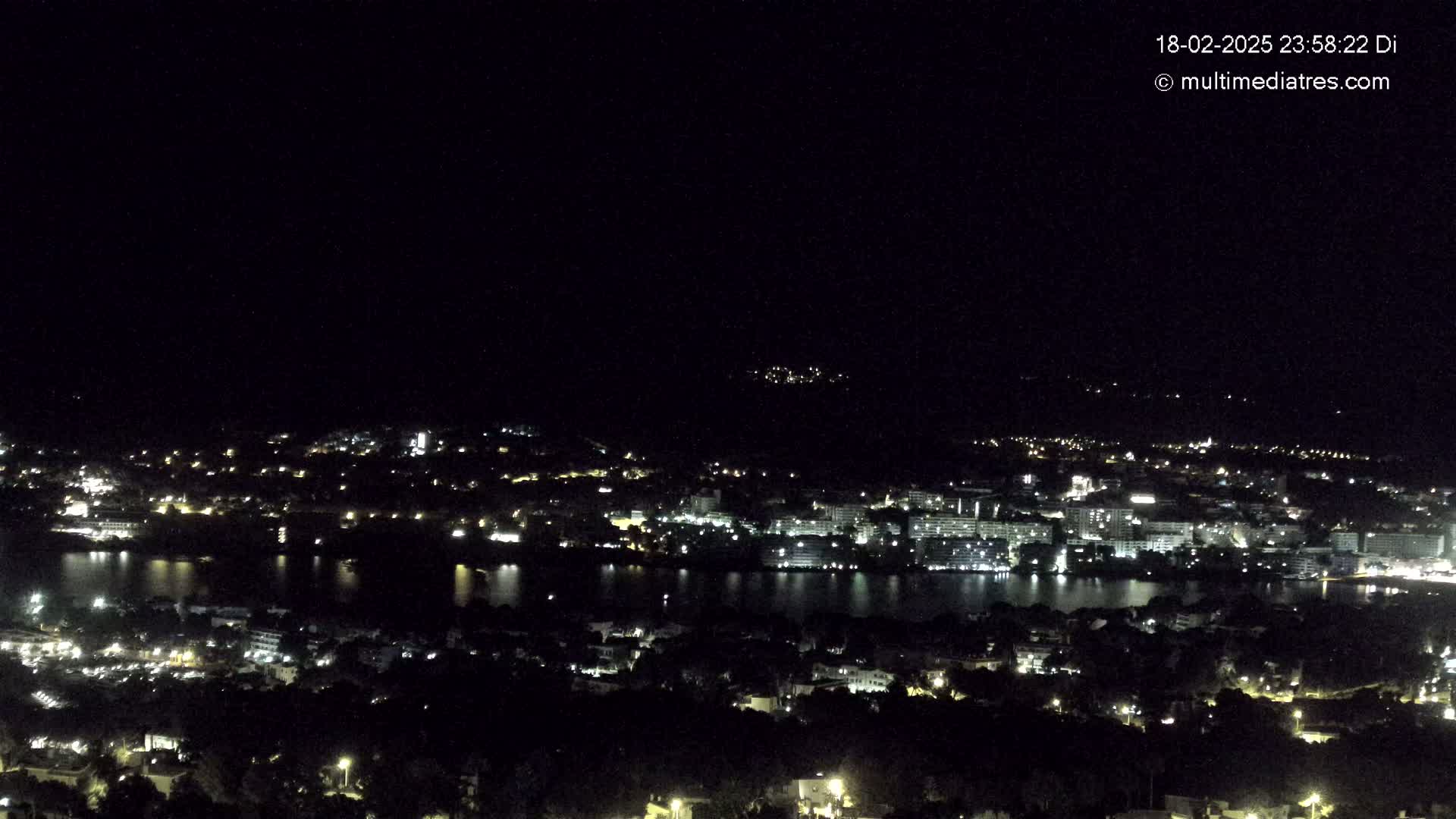 A nighttime, high-angle view shows a city with numerous lights surrounding a dark body of water under a clear, dark sky.