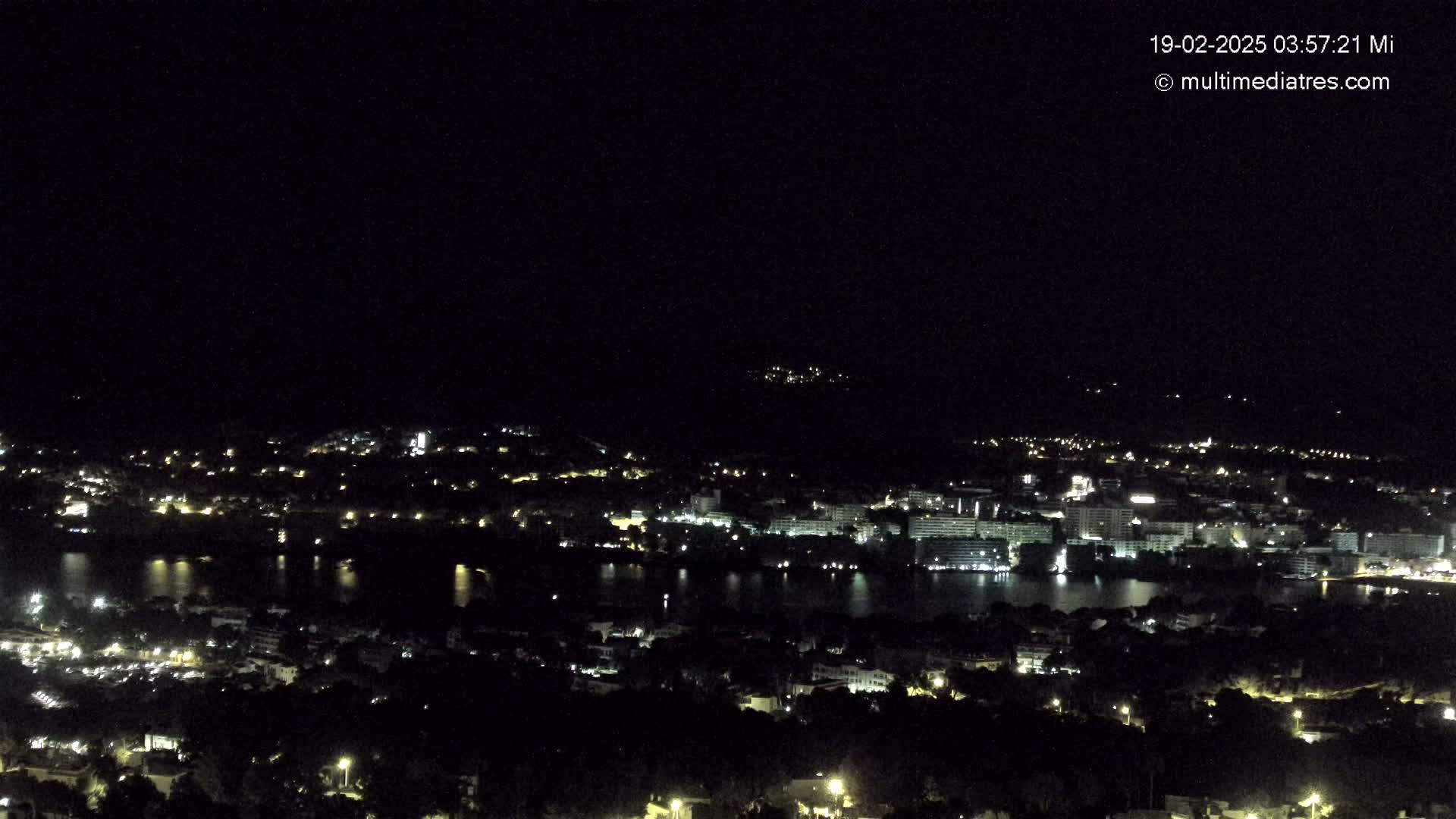 A nighttime, aerial view of a coastal city with its lights illuminated against a dark sky, reflecting in a body of water.