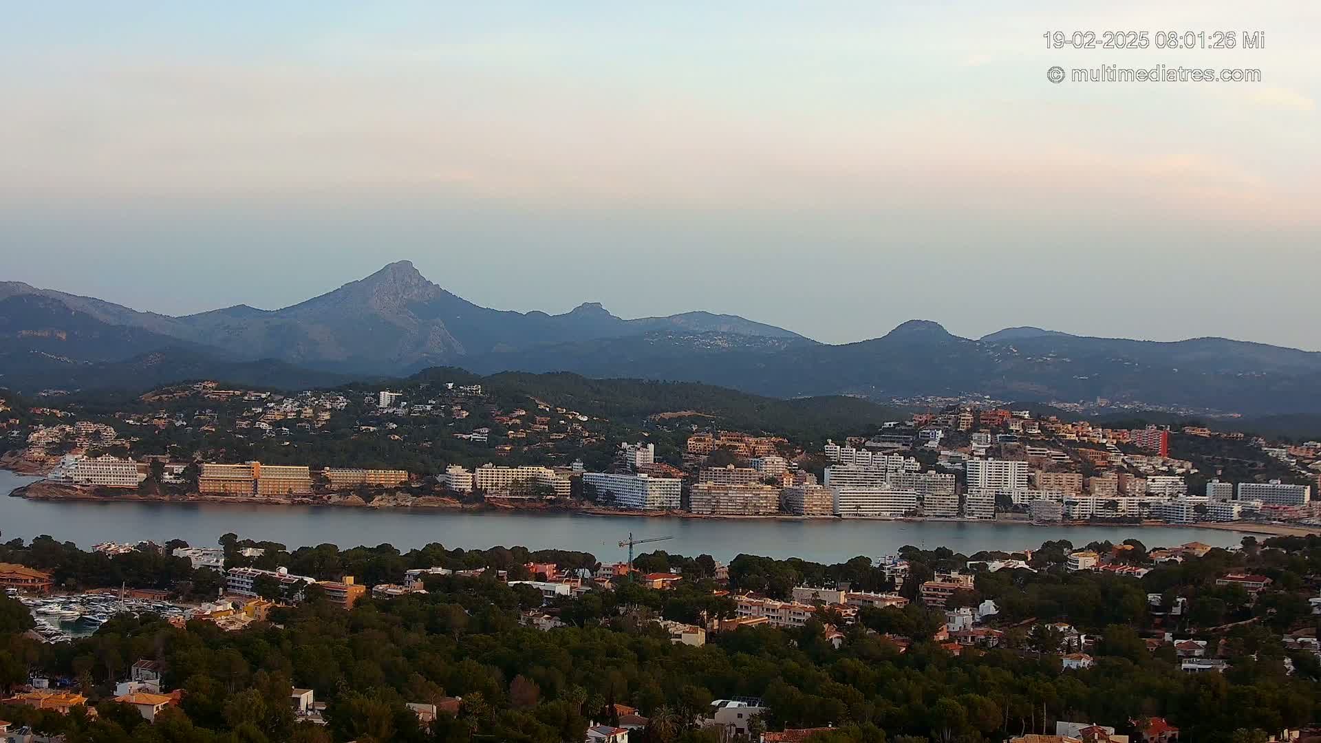 A coastal town nestled between a calm bay and a mountain range under a pale, clear sky.
