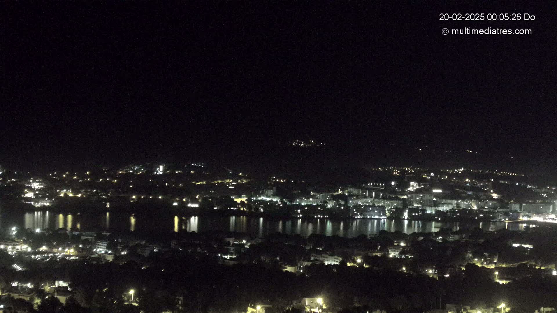 A nighttime view of a city illuminated by lights, nestled beside a calm body of water reflecting the city's glow under a clear, dark sky.