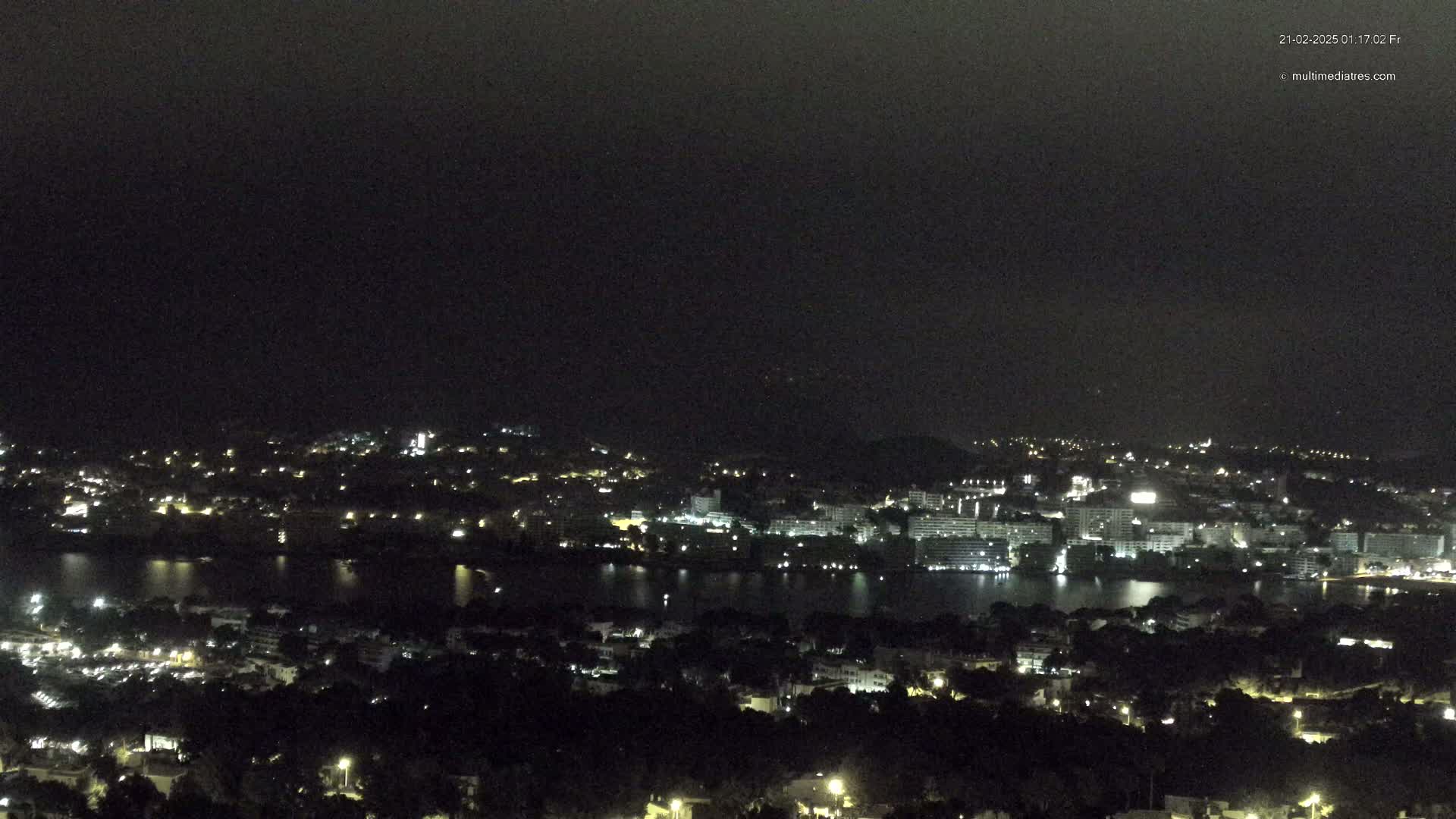 A nighttime, long-range view of a city illuminated by lights, situated alongside a dark body of water, under a mostly overcast sky.
