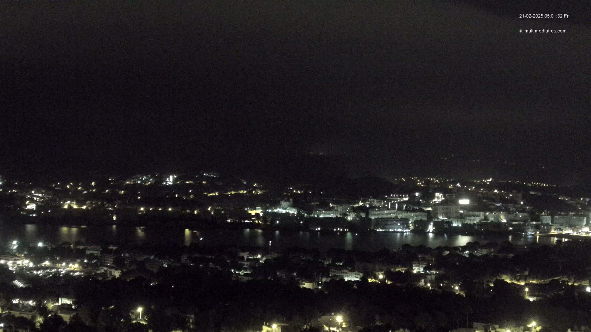 A nighttime, high-angle view of a city illuminated by lights surrounding a dark body of water, under a mostly cloudy sky.