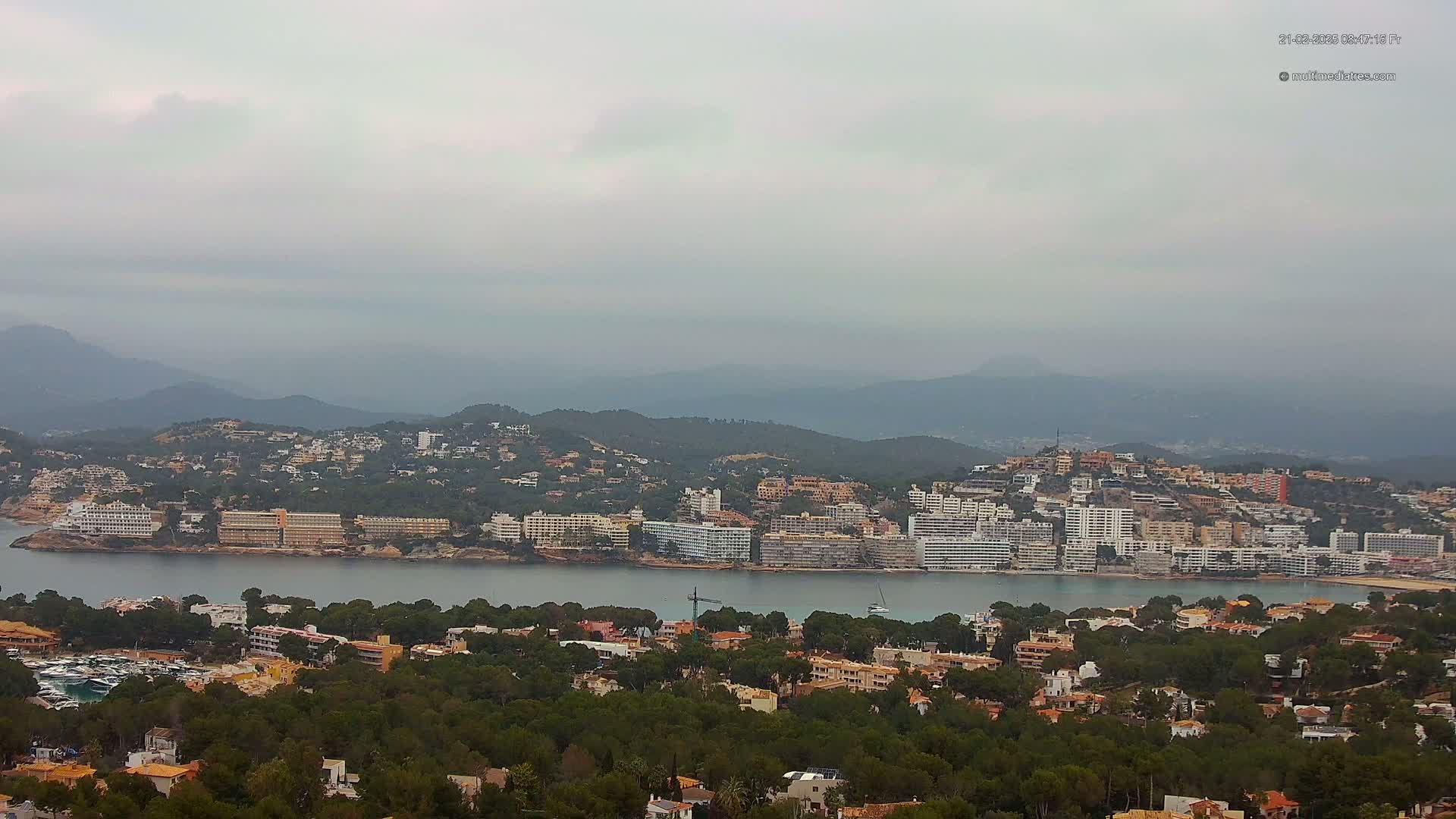 A coastal town nestled in a bay, surrounded by lush greenery and hazy mountains under an overcast sky.