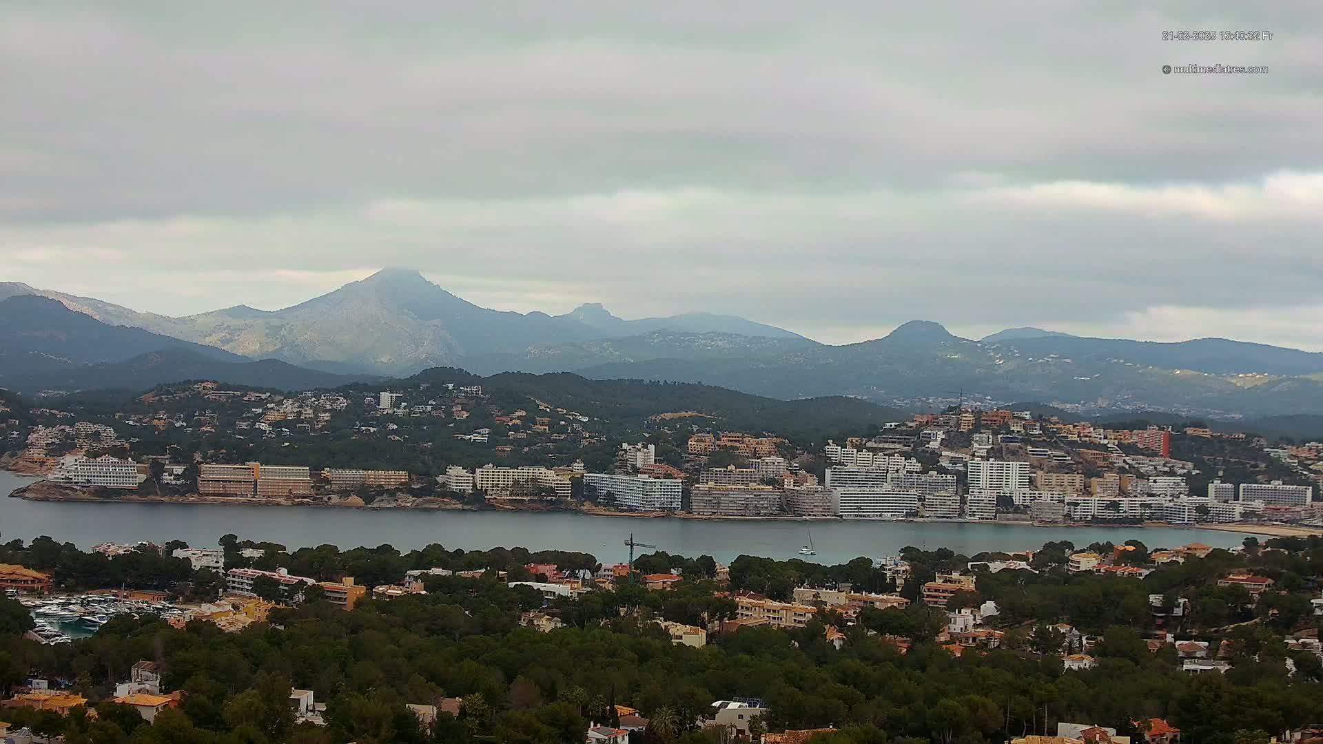 A coastal town nestled between a calm bay and a mountain range under an overcast sky.