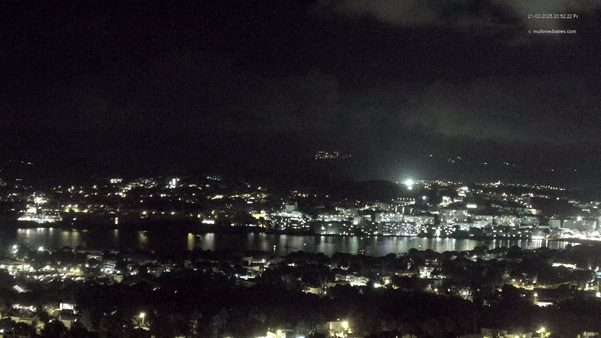 A nighttime view of a city illuminated by lights, situated along a body of water under a cloudy sky.