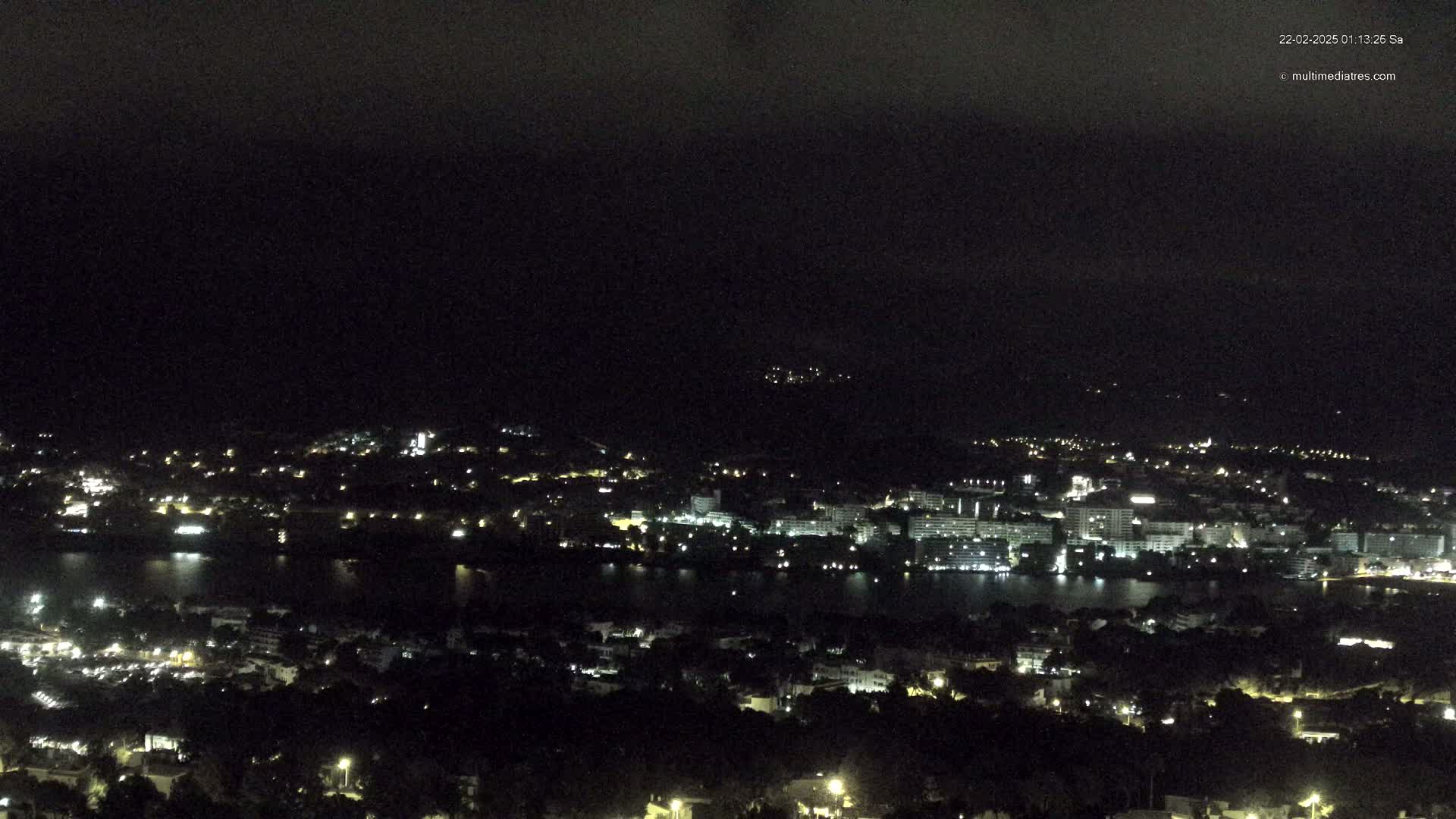 A nighttime, high-angle view shows a city with many lights bordering a dark body of water, under a dark, cloudy sky.