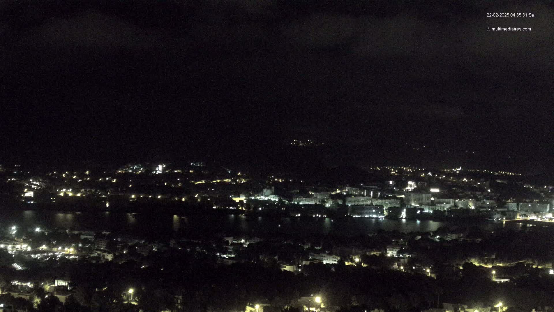 A nighttime, high-angle view shows a coastal city with scattered lights illuminated against a dark, cloudy sky.