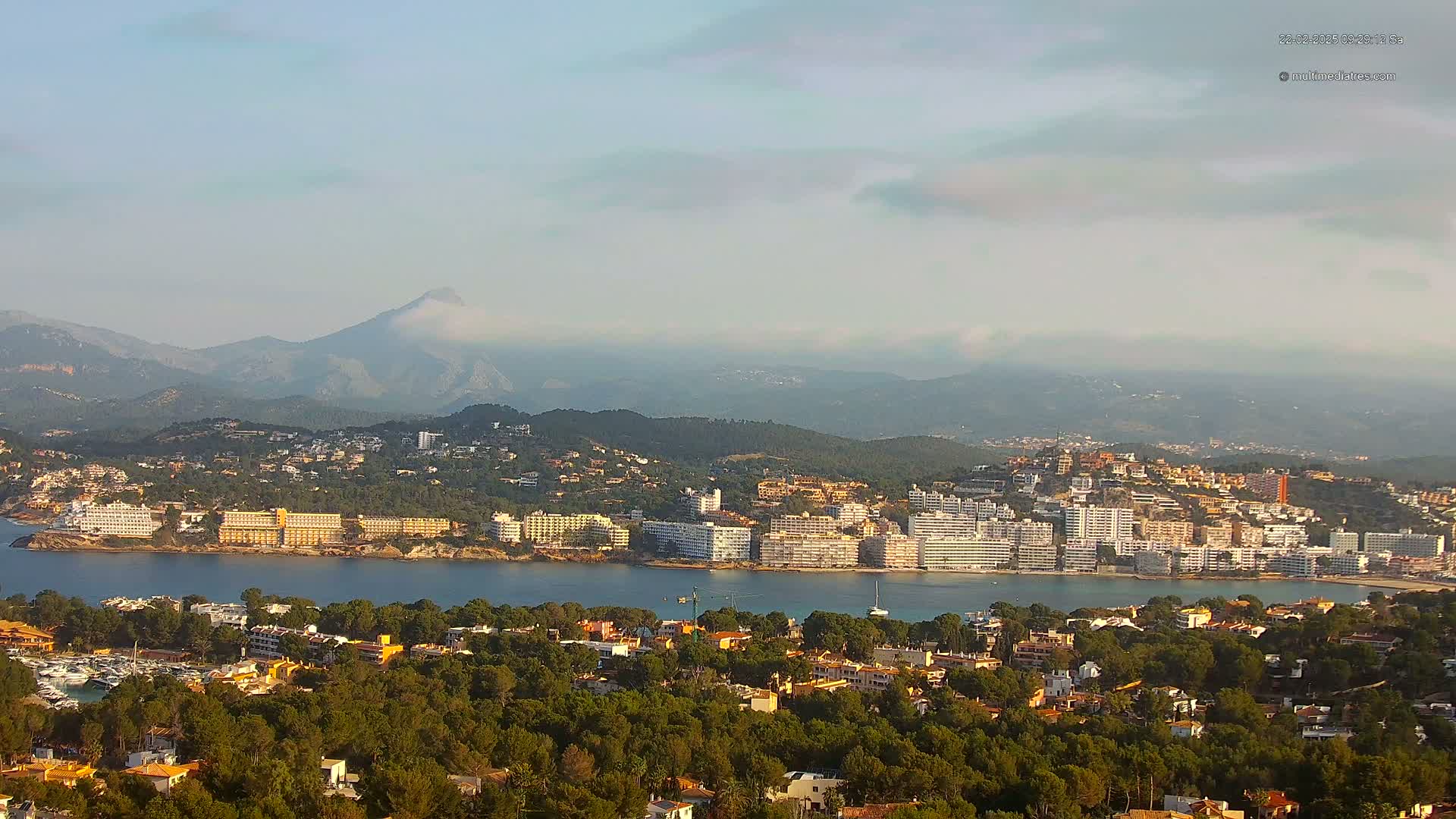 A coastal town nestled among green hills and mountains under a hazy sky, with a body of water separating the town from a line of hotels.