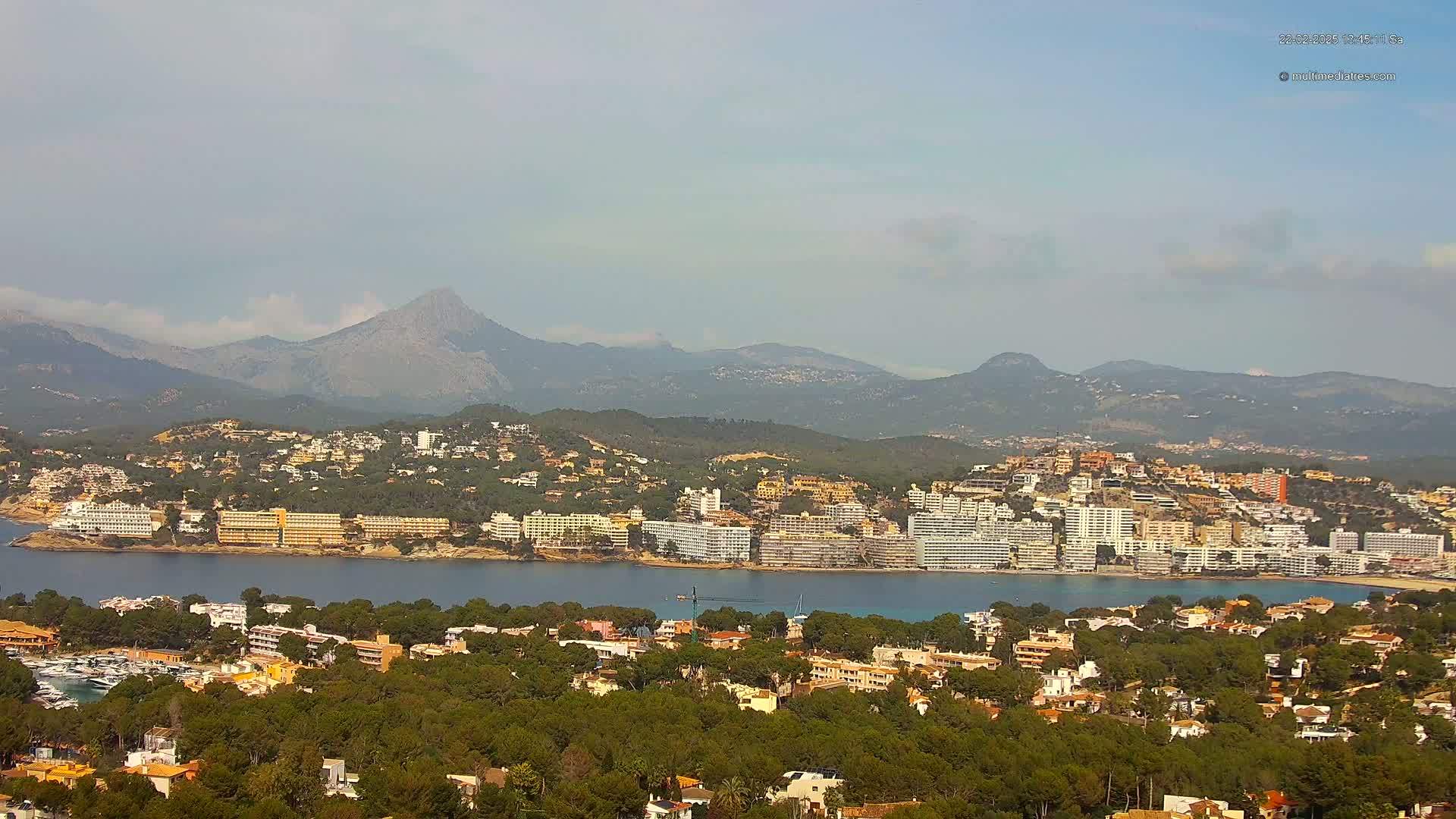 A coastal town nestled amongst green hills and mountains, overlooks a calm bay under a partly cloudy sky.