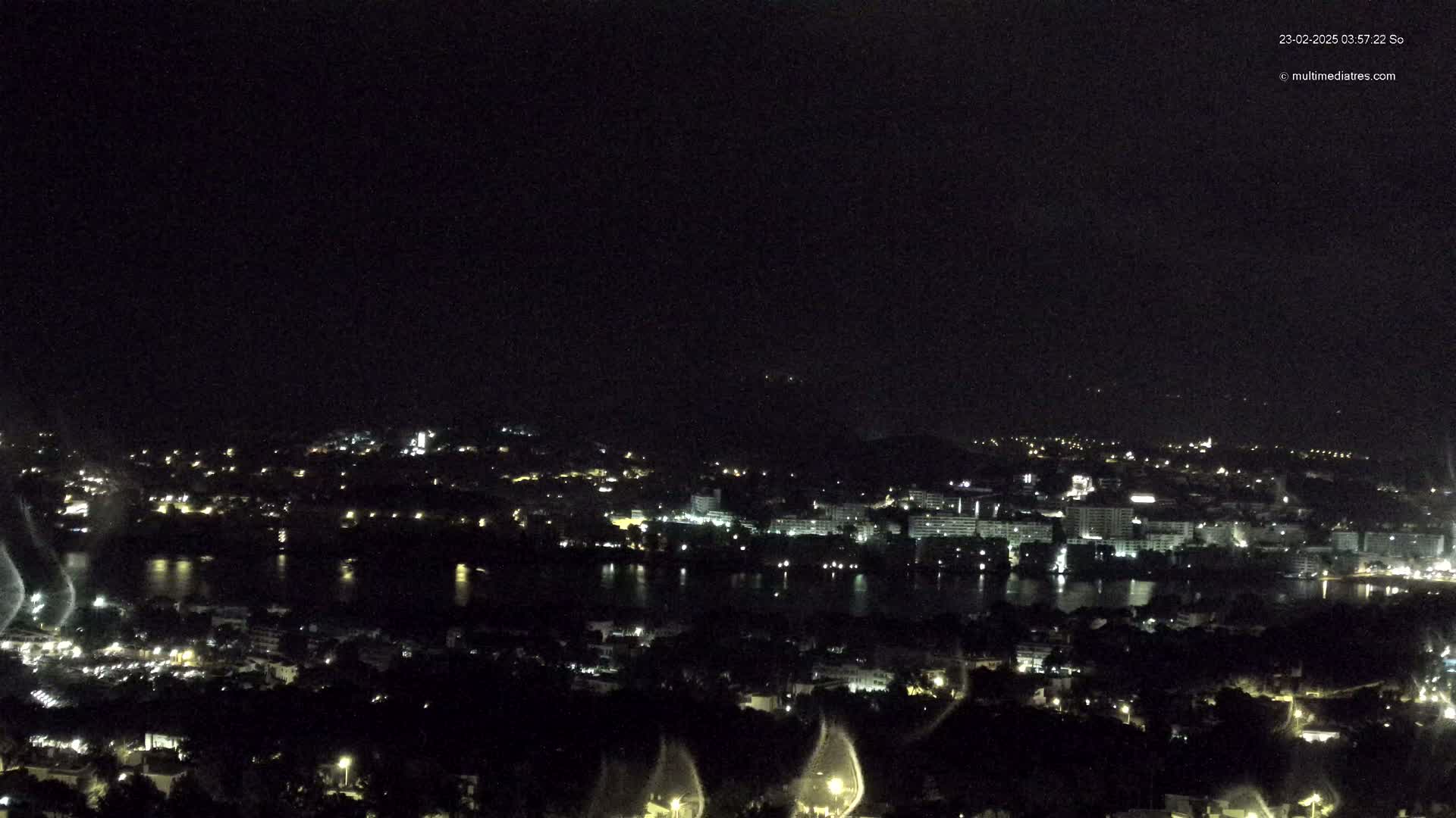 A nighttime, high-angle view of a city with a body of water, possibly a lake or bay, in the foreground, under a dark, clear sky.