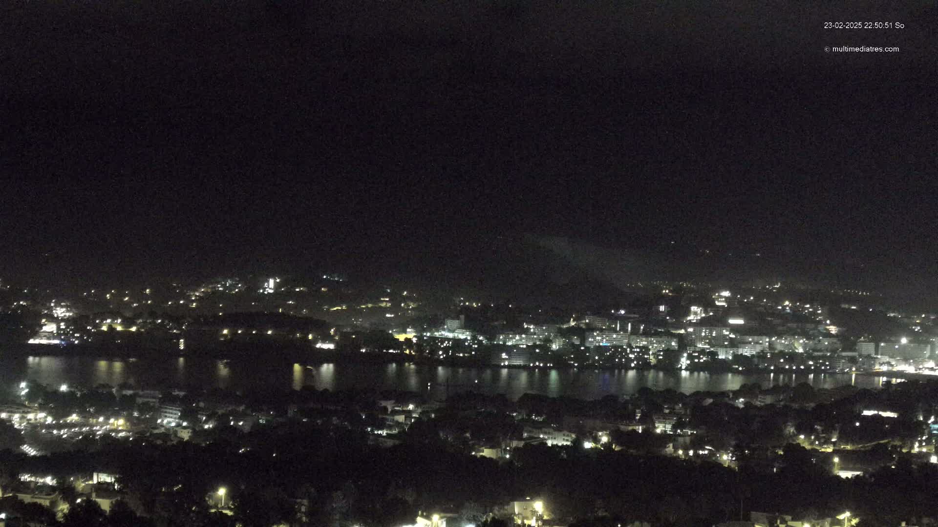 A nighttime view of a city situated on a body of water, under a dark, cloudy sky.