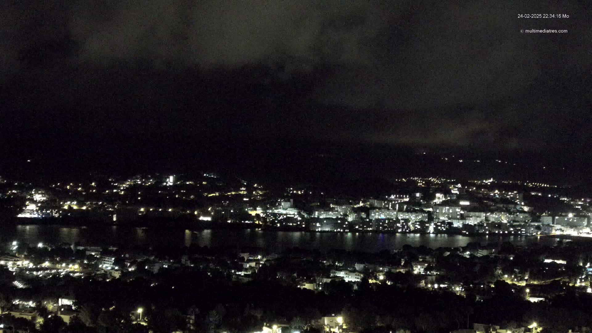 A nighttime, high-angle view of a city sprawling across a body of water under a dark, cloudy sky.