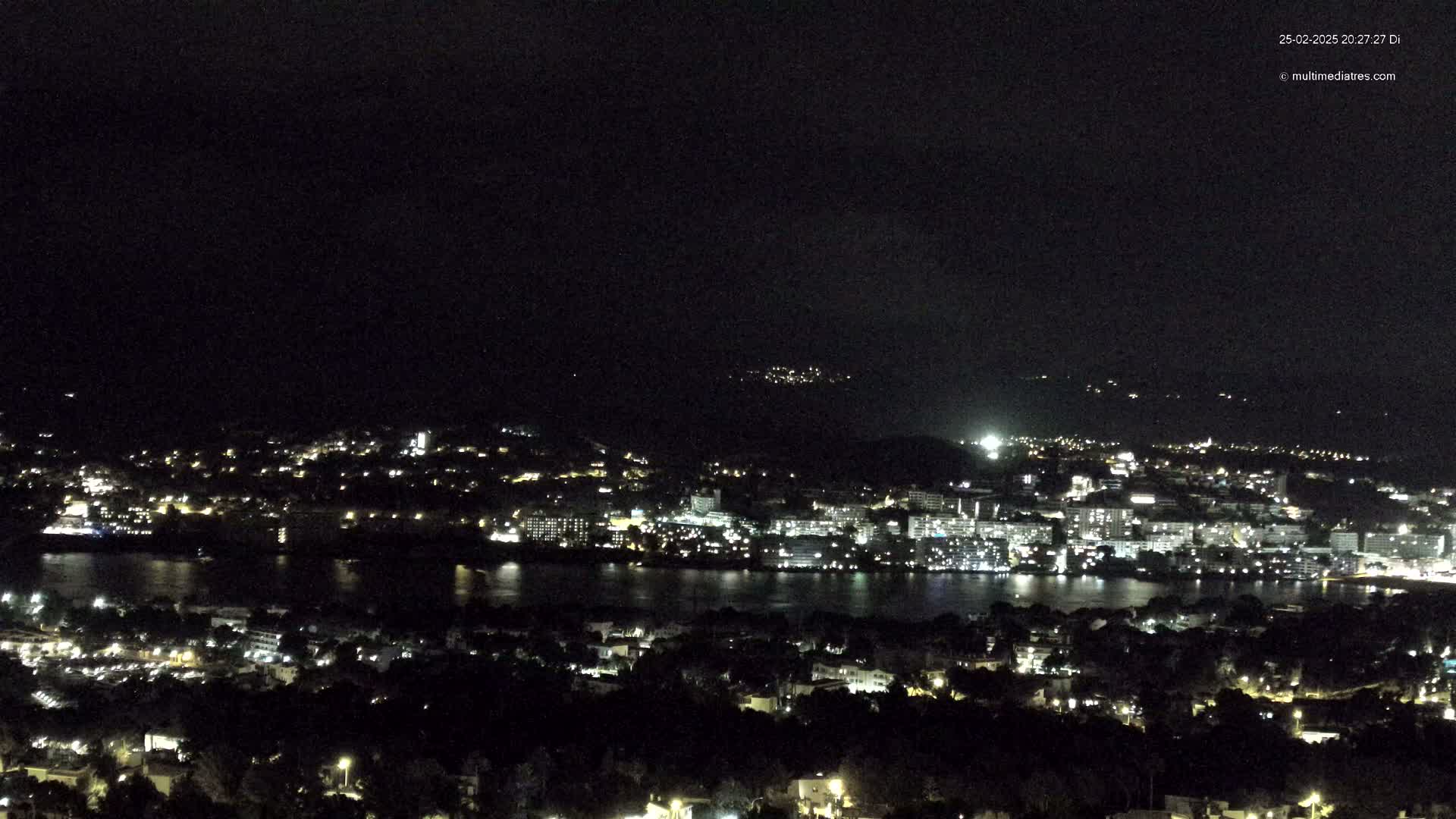 A nighttime, aerial view of a coastal city illuminated with lights, across a dark body of water, under a clear, dark sky.