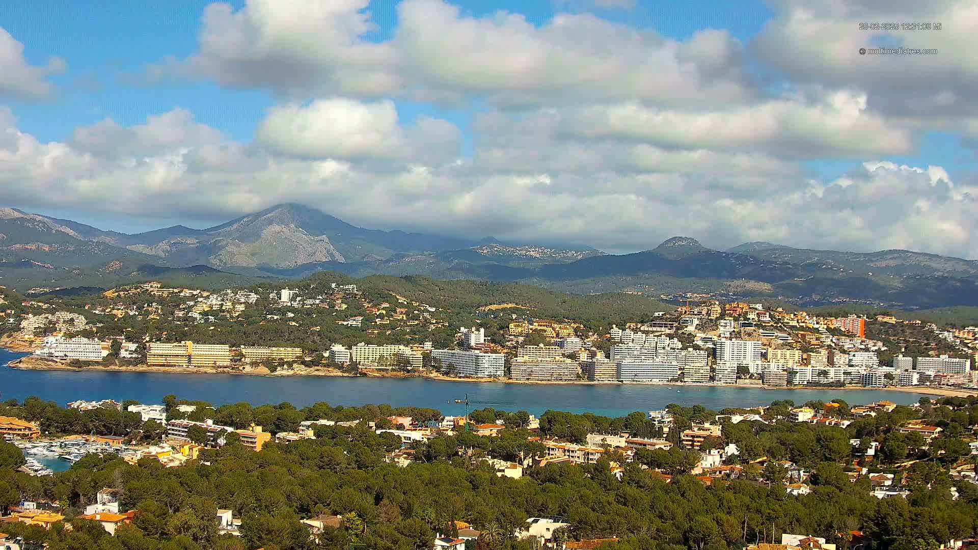 A coastal town nestled between a bay and a mountain range under a partly cloudy sky.