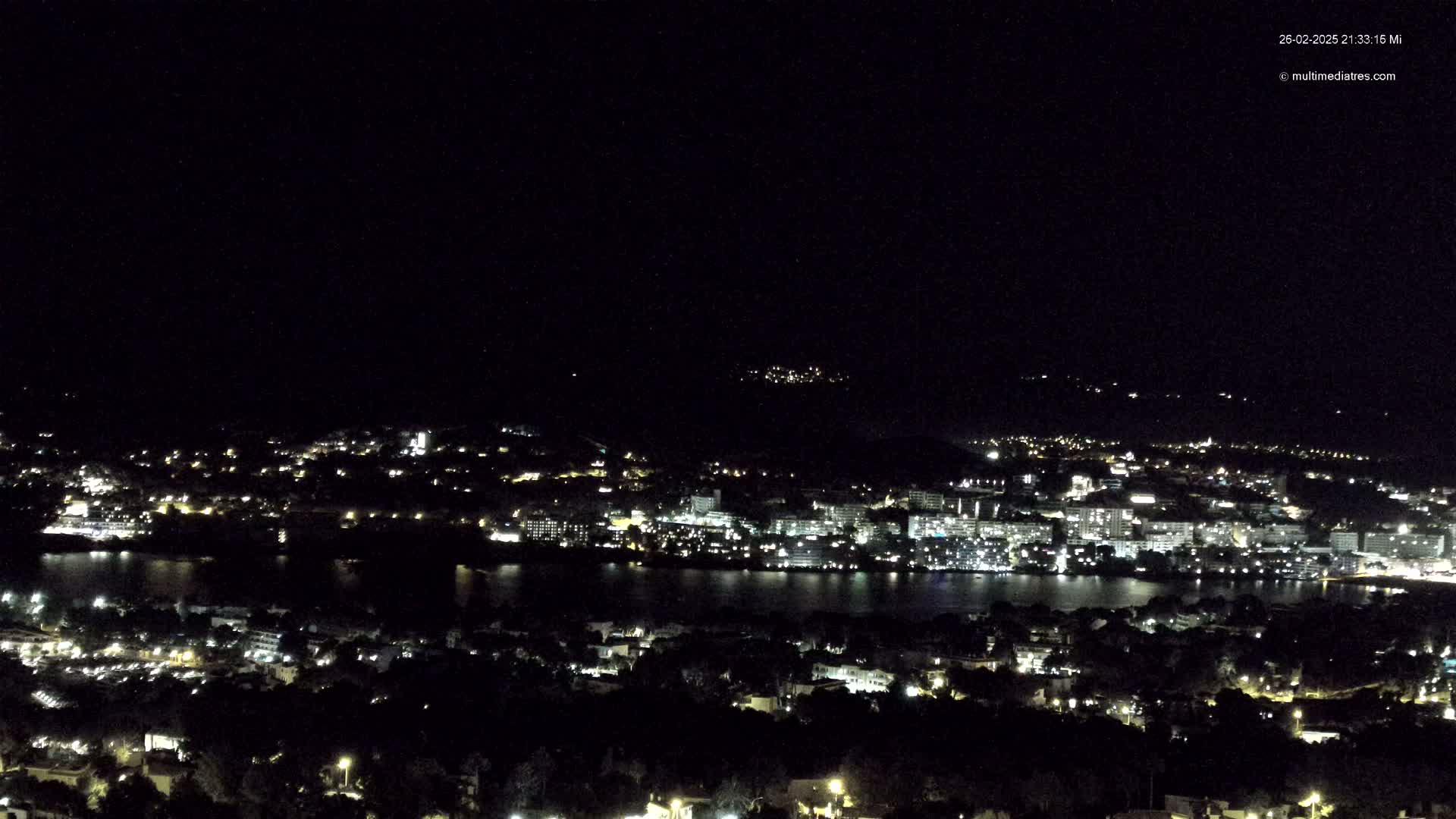 A nighttime, long-range view of a coastal city illuminated by numerous lights, situated beside a dark body of water.