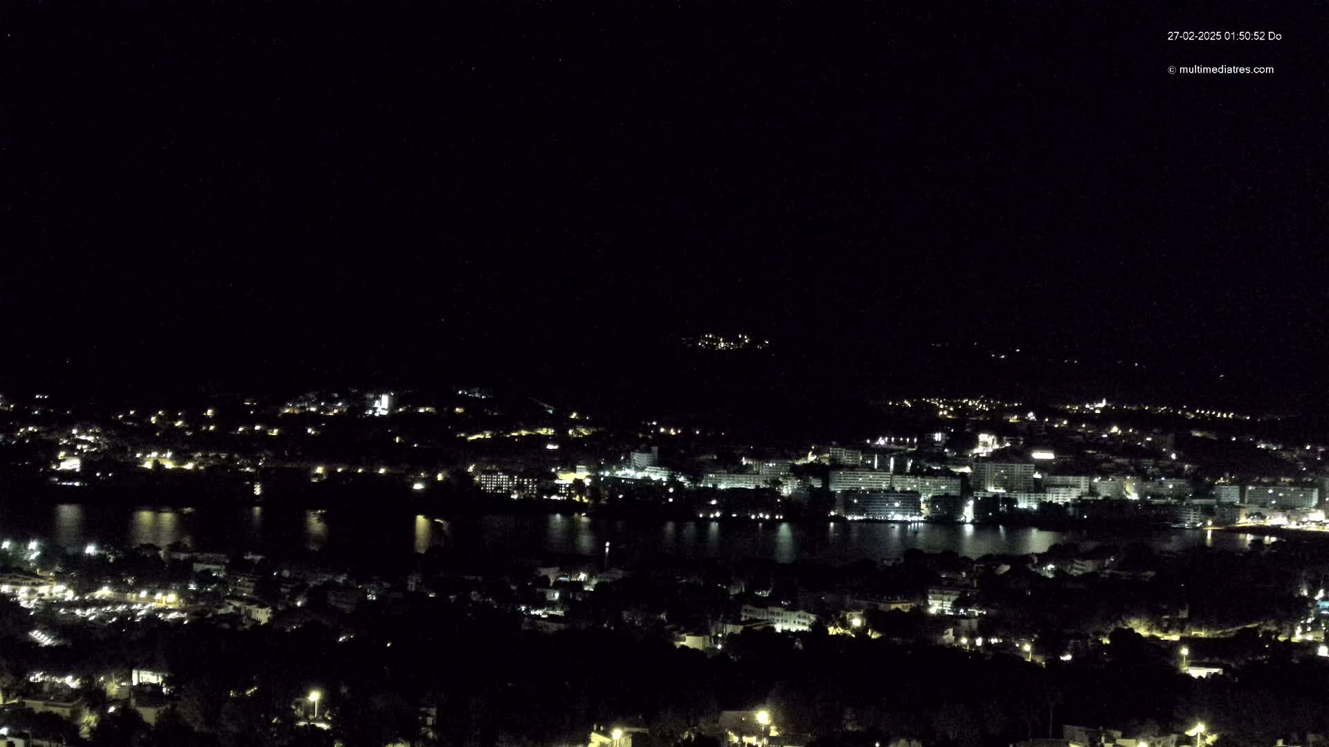 A nighttime, high-angle view of a coastal city with a body of water, featuring numerous lights illuminating buildings and streets under a clear, dark sky.
