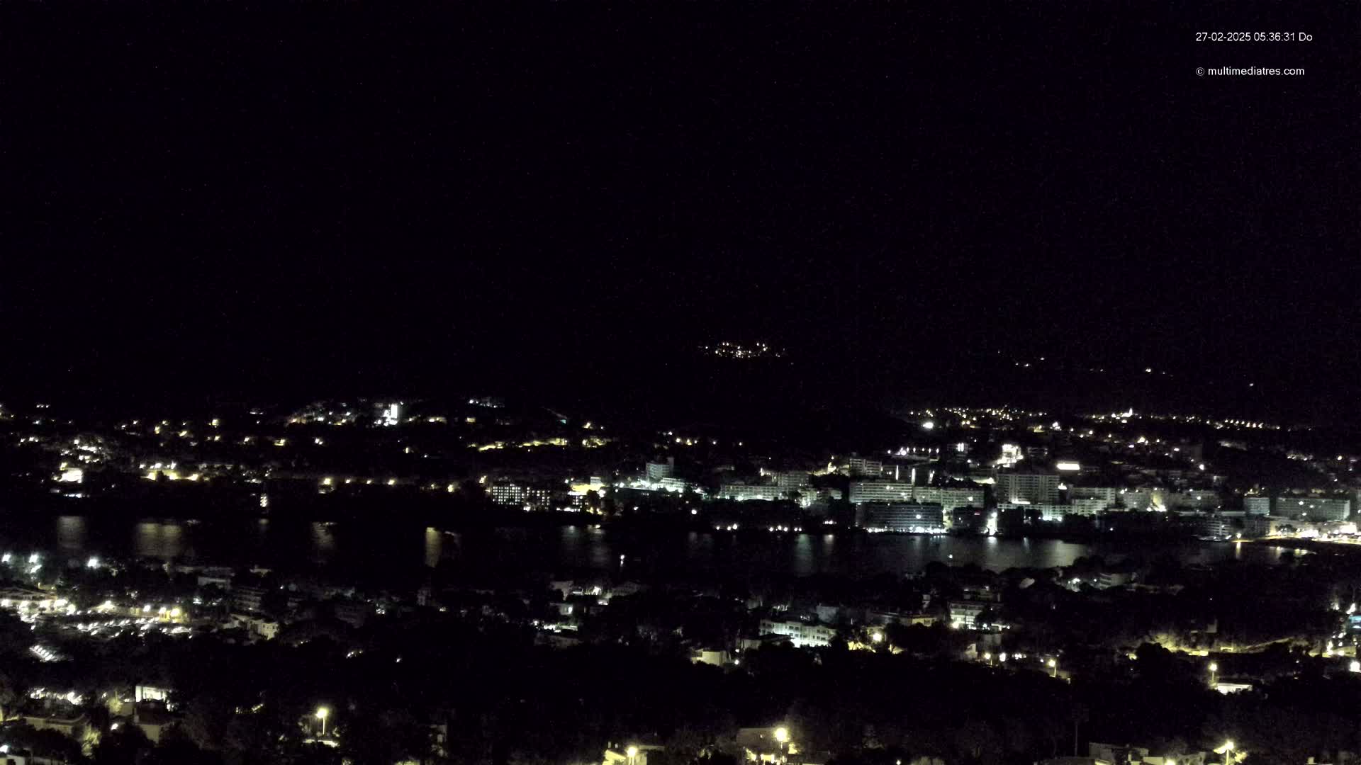 A nighttime, high-angle view shows a coastal town with many lights illuminating buildings and streets, next to a dark body of water, under a clear, dark sky.