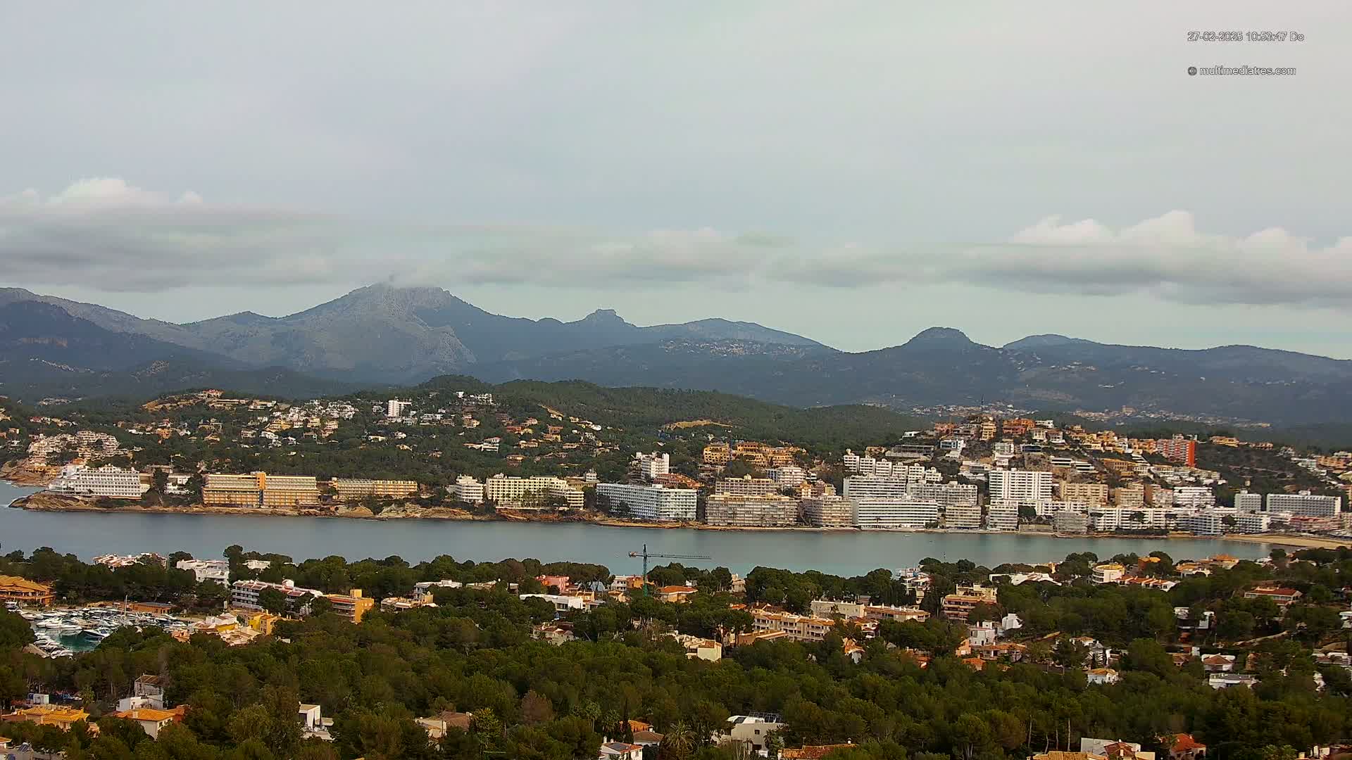 A coastal town with many hotels and buildings sits nestled between a calm bay and a mountain range under a mostly cloudy sky.
