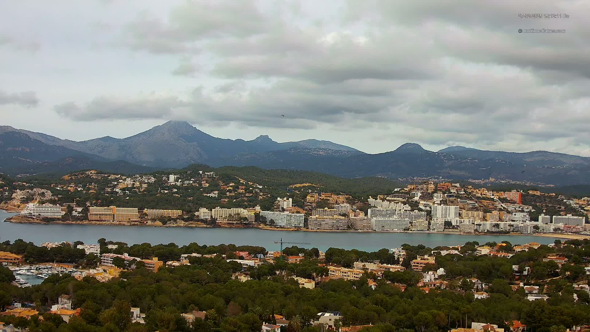 A coastal town with numerous buildings stretches along a calm bay, backed by mountains under a cloudy sky.