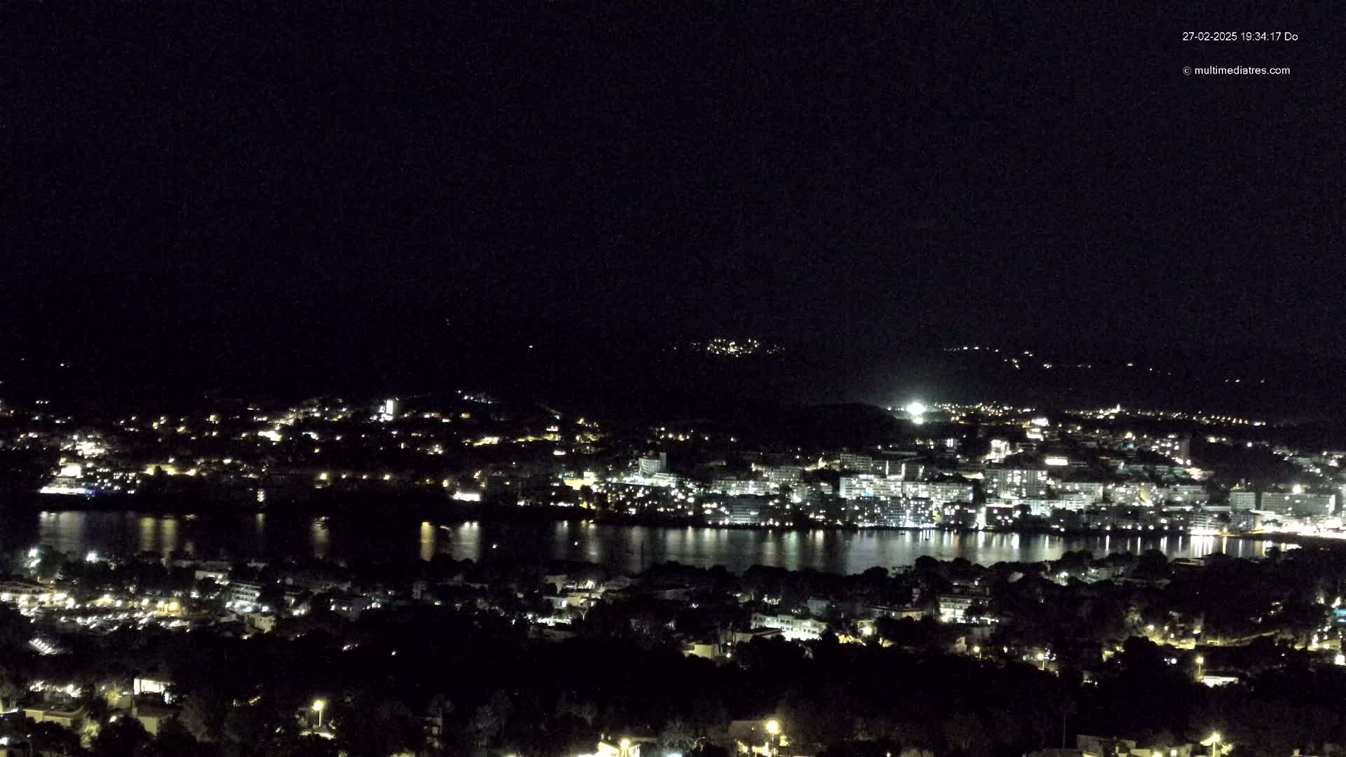 A nighttime, high-angle view of a coastal city illuminated by lights, with calm water reflecting the city lights.