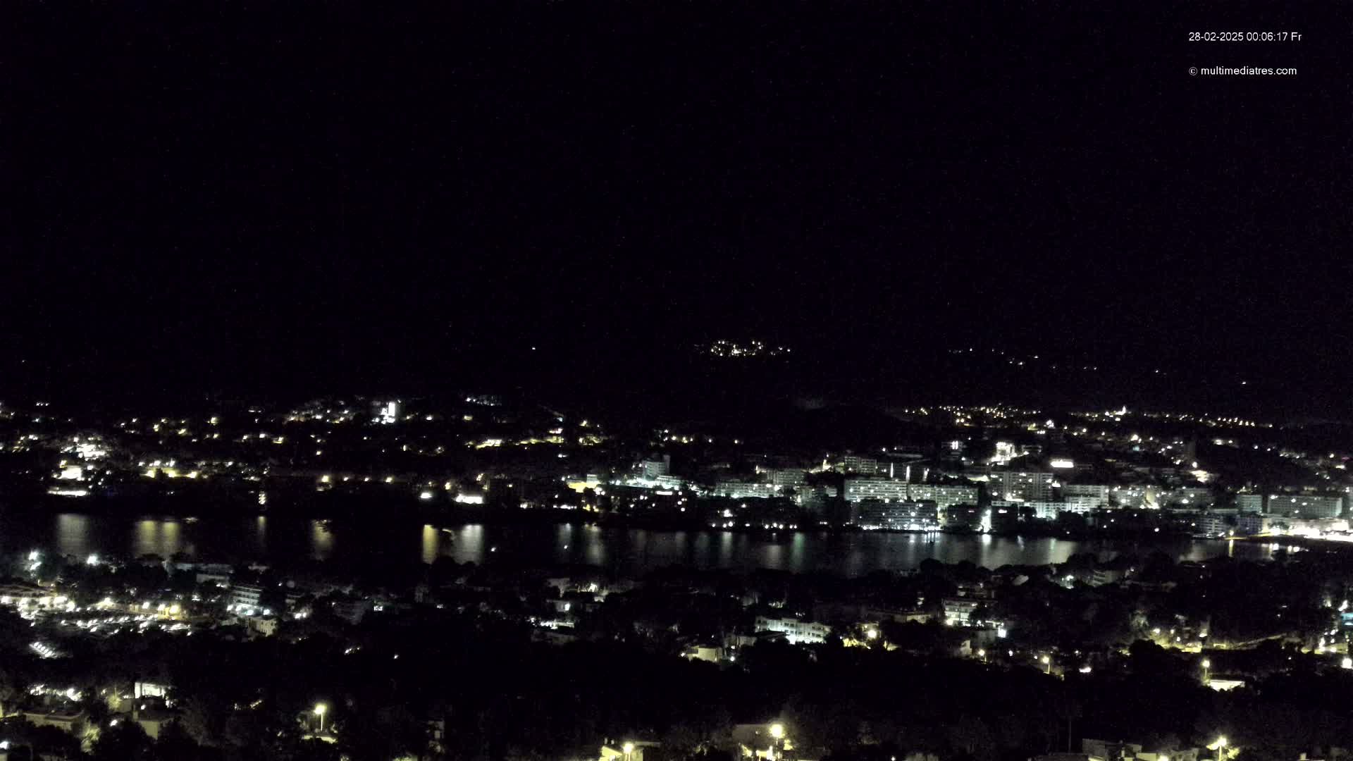 A nighttime, high-angle view shows a city with lights illuminated near a body of dark water, under a clear, dark sky.