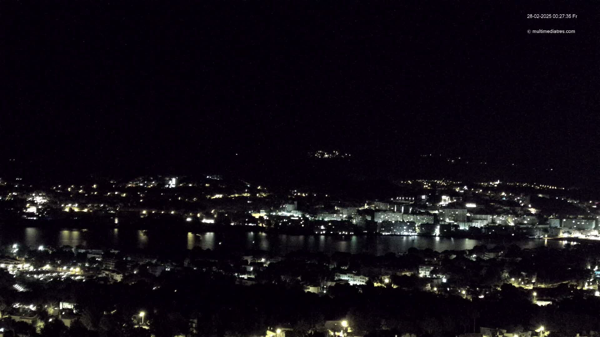 A nighttime view of a coastal city illuminated by scattered lights, with a calm body of water separating the city from a hilltop vantage point.