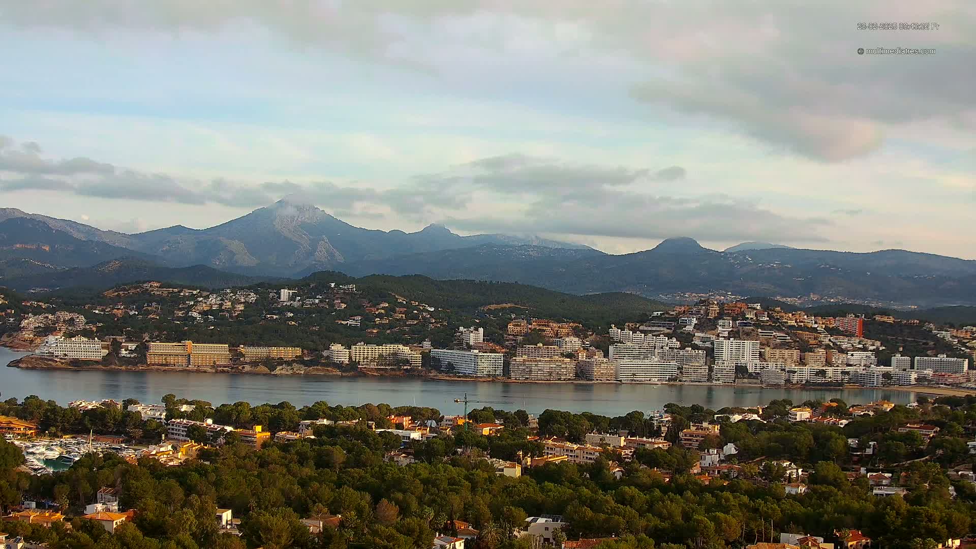 A coastal town nestled between a calm bay and a mountain range under a partly cloudy sky.