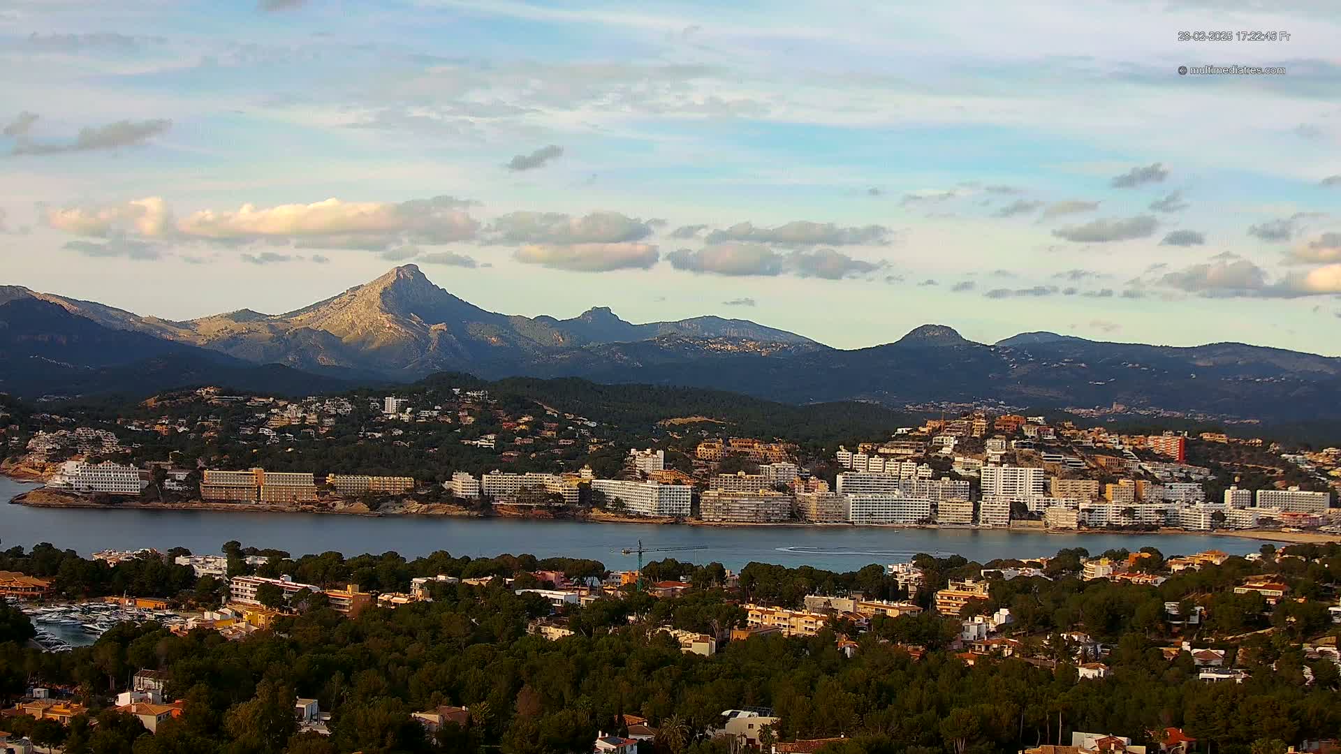A coastal town with numerous buildings sits beside a calm bay, backed by a range of mountains under a partly cloudy sky.