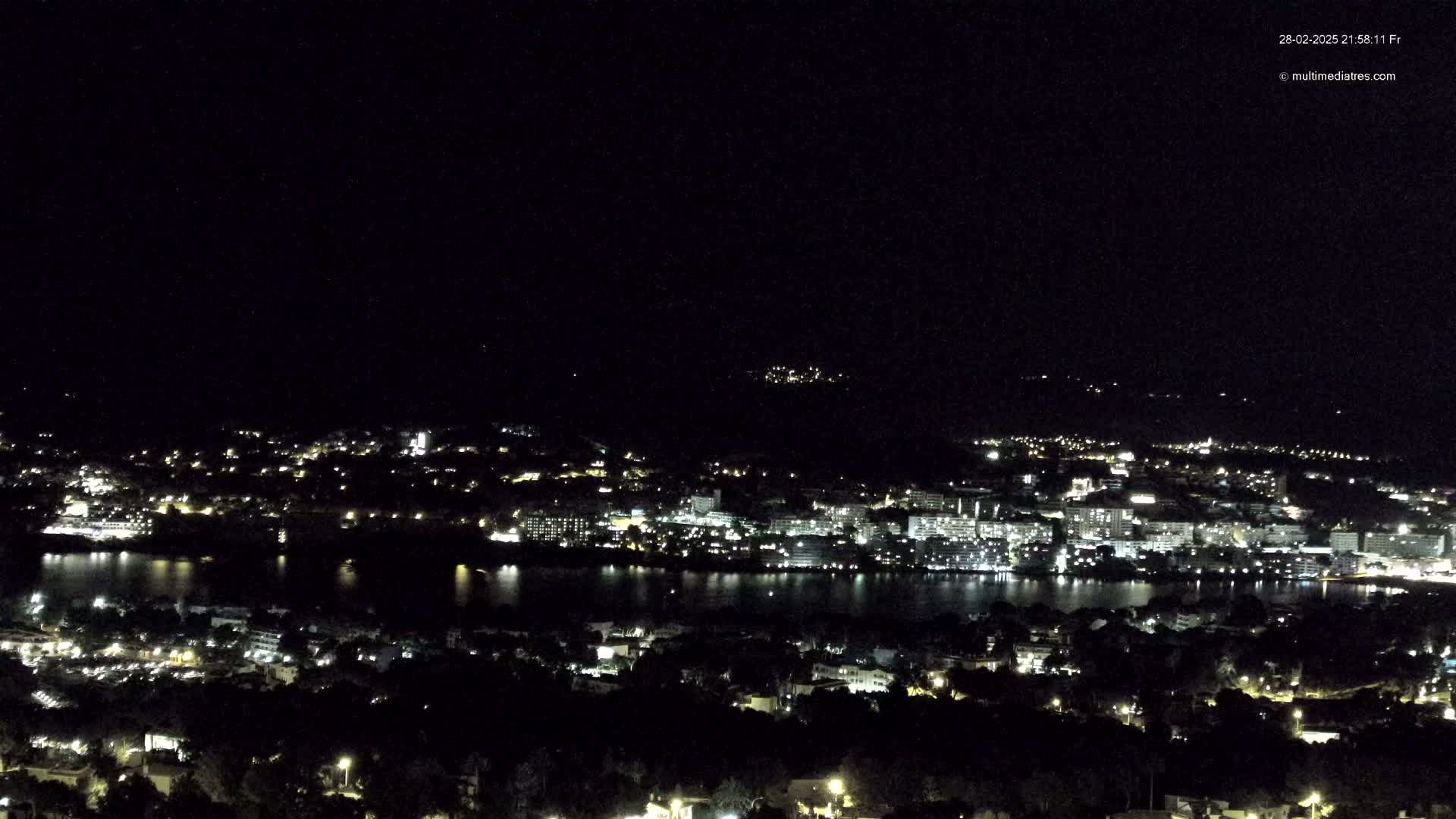 A nighttime view of a city illuminated by lights, situated beside a body of dark water, under a clear, dark sky.