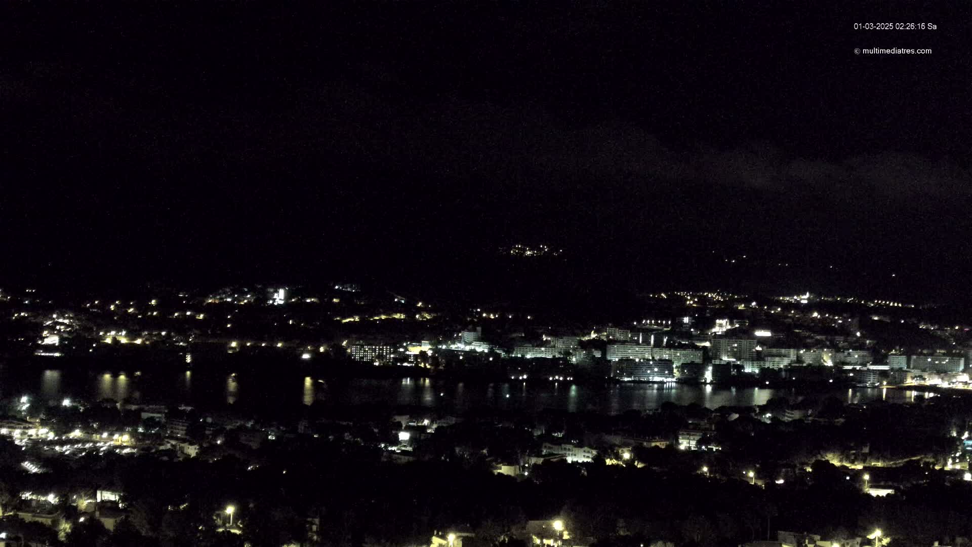 A nighttime, high-angle view of a coastal city with a body of water reflecting the lights, under a dark, cloudy sky.
