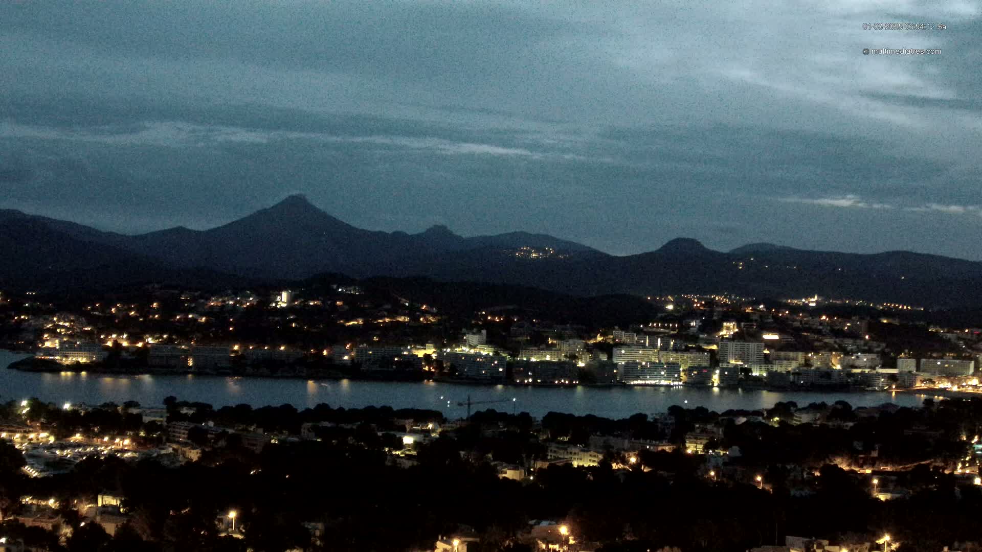 A nighttime view of a coastal town illuminated with lights, nestled between a dark body of water and a range of silhouetted mountains under a dusky sky.