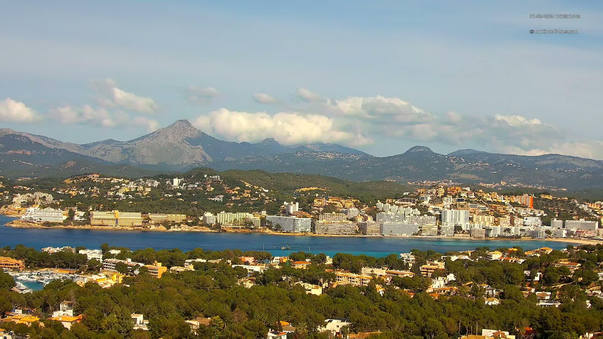 A coastal town nestled among green hills and mountains under a partly cloudy sky, with a calm body of water separating the town from a line of hotels.