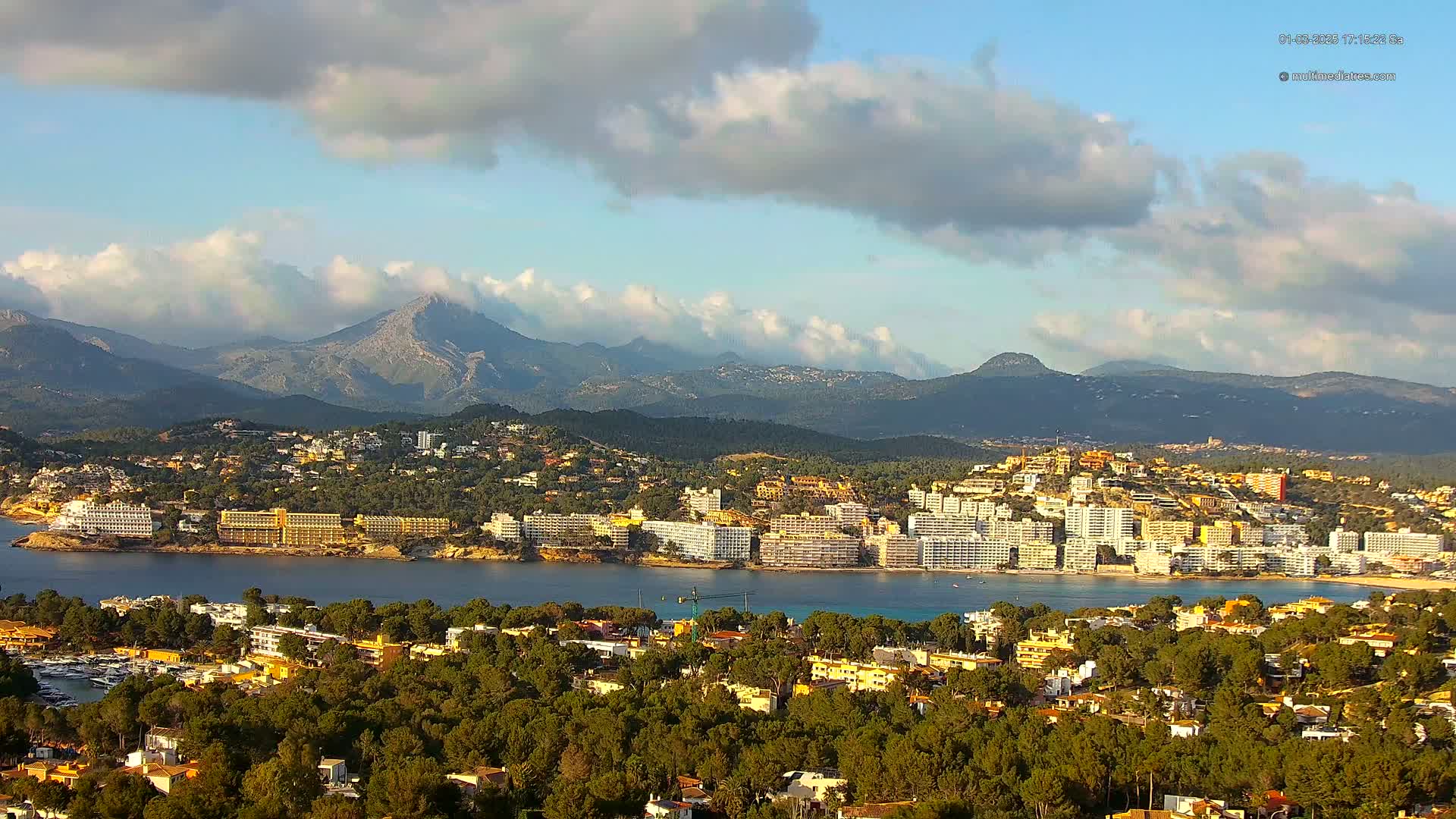 A coastal town with numerous hotels and buildings is nestled at the base of a mountain range under a partly cloudy sky.