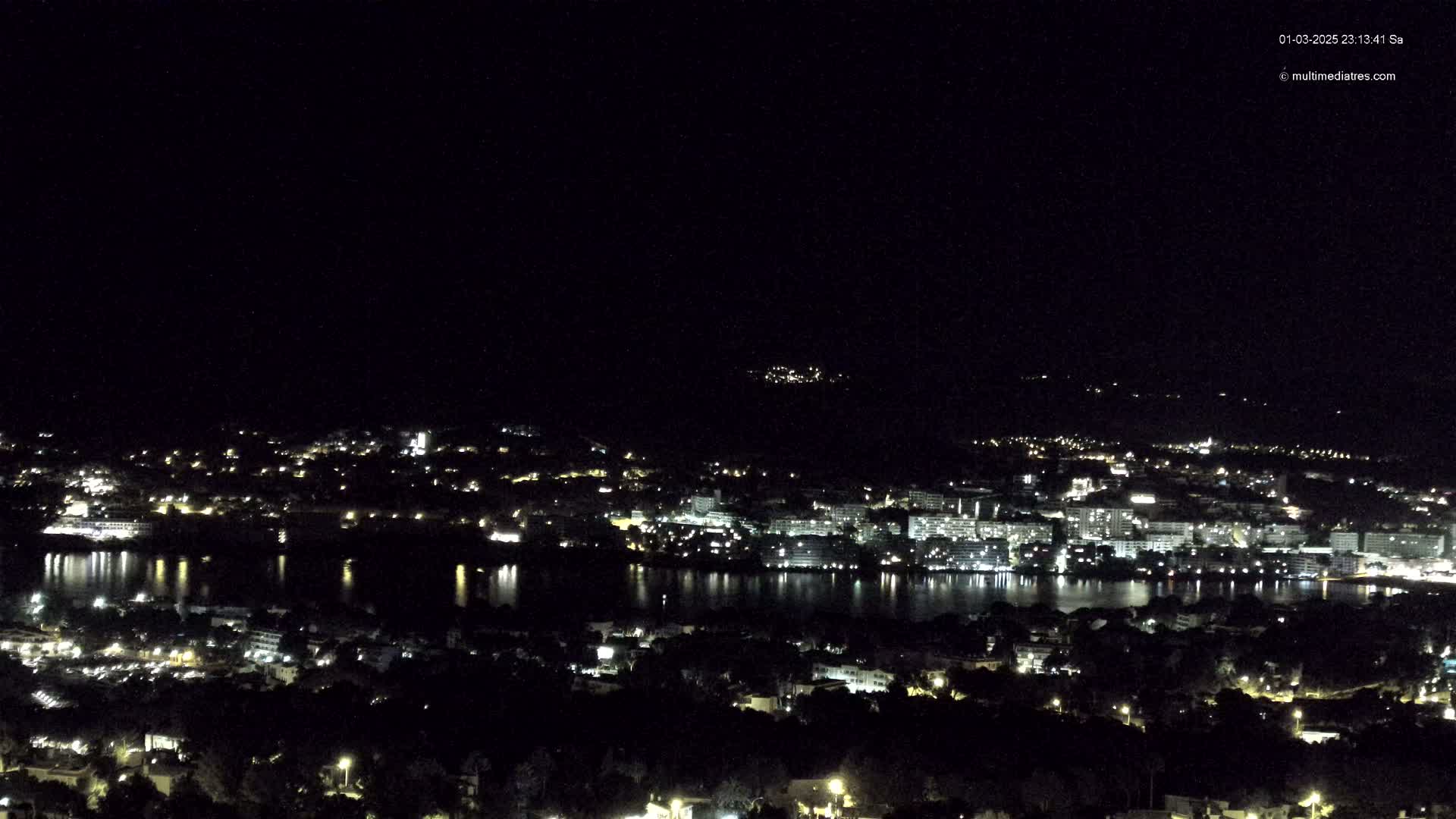 A nighttime, high-angle view shows a city with lights reflecting on a body of water.