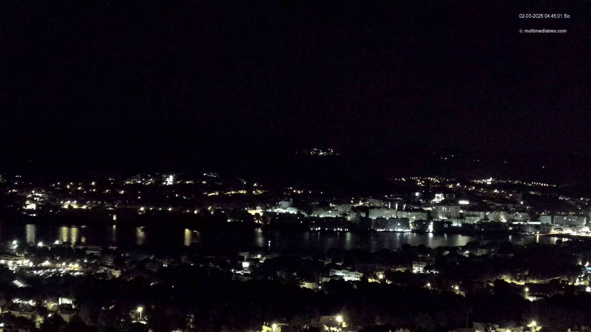 A nighttime view of a coastal town with a body of water reflecting the lights, under a clear, dark sky.