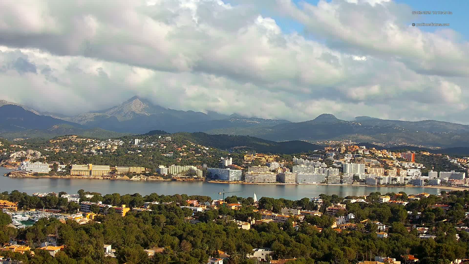 A coastal town nestled between a calm bay and a mountain range under a partly cloudy sky.